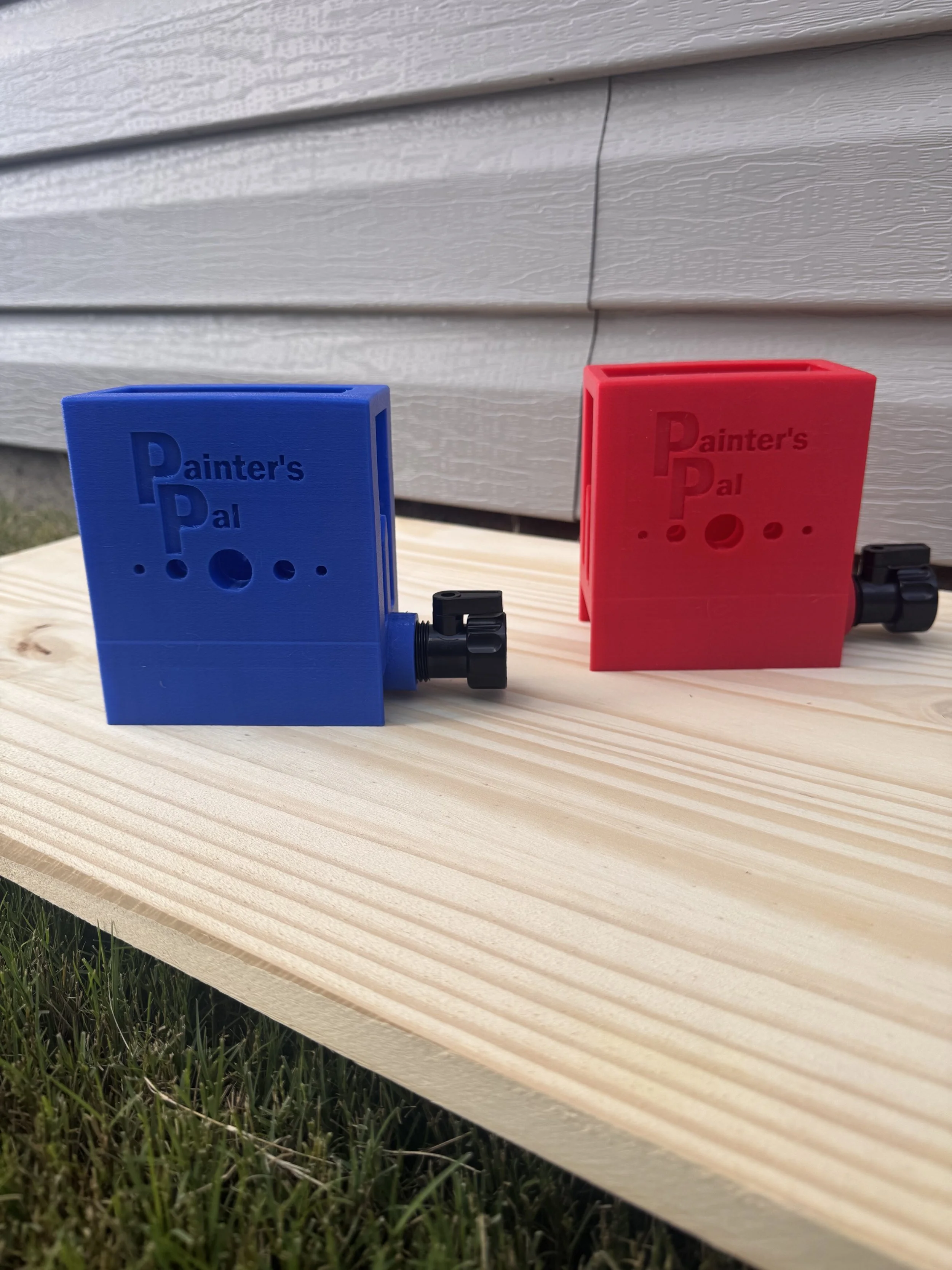 Two paintbrush cleaning attachment tools, one blue and one red, with black adjustment knobs, placed on a wooden surface outdoors next to a house with horizontal siding.