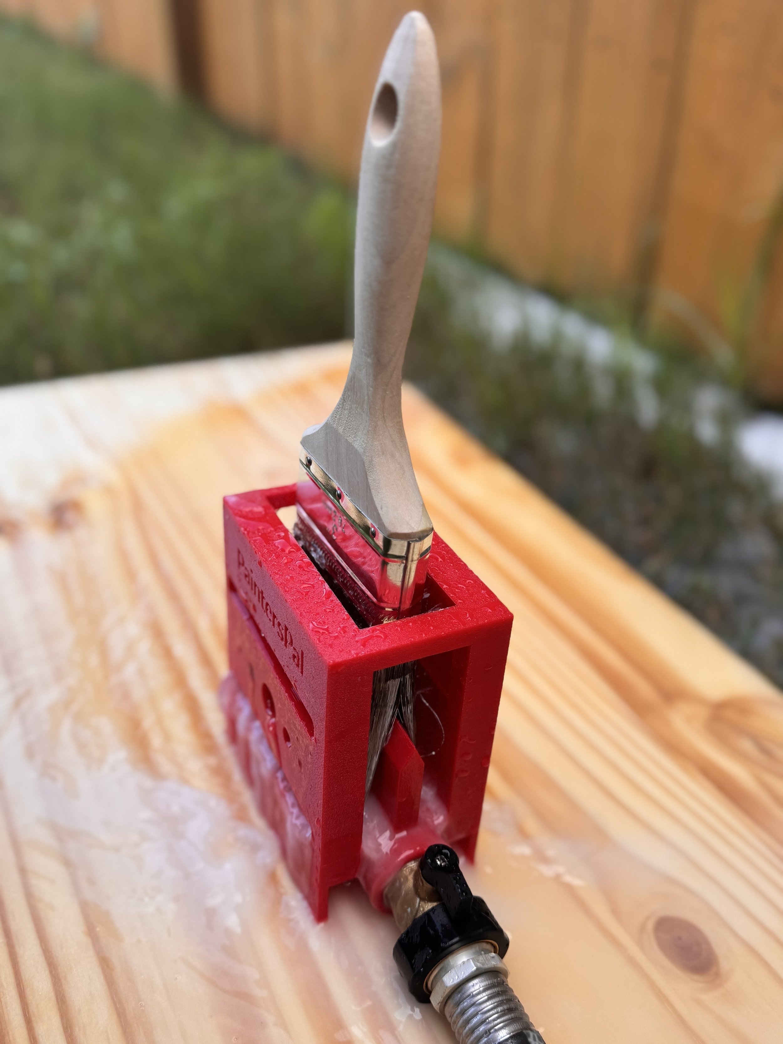 A small paintbrush cleaning tool in red, attached to a water supply, placed on a wooden surface outdoors.