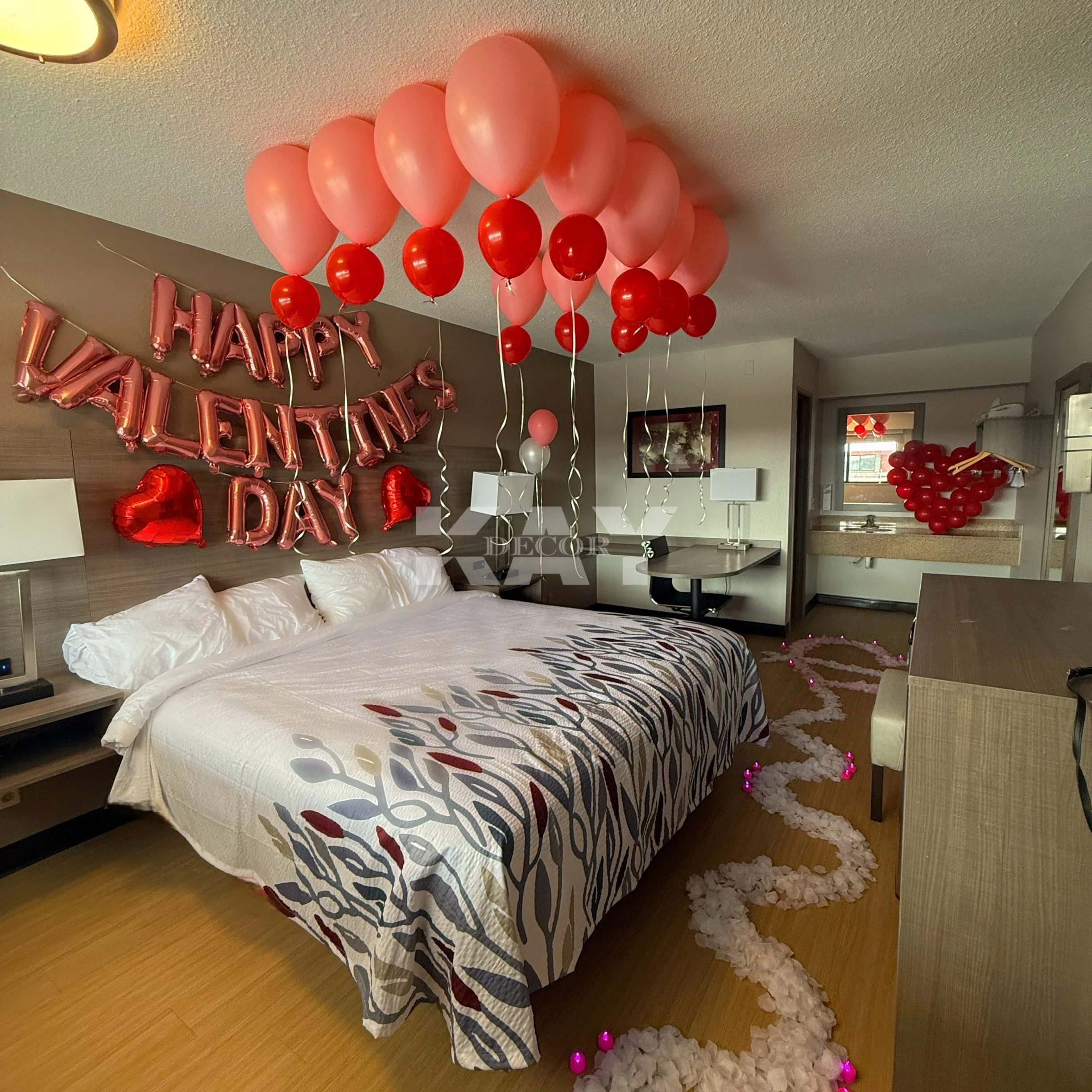 A hotel room decorated for Valentine's Day with pink and red balloons hanging from the ceiling. A pink 'Happy Valentine's Day' balloon banner is on the wall behind the bed, which has white pillows and a patterned bedspread. Red balloons are attached to the balloons hanging from the ceiling. A pink and white flower petal trail winds across the floor, illuminated with small pink lights, leading toward a window with additional red balloons arranged in a heart shape.