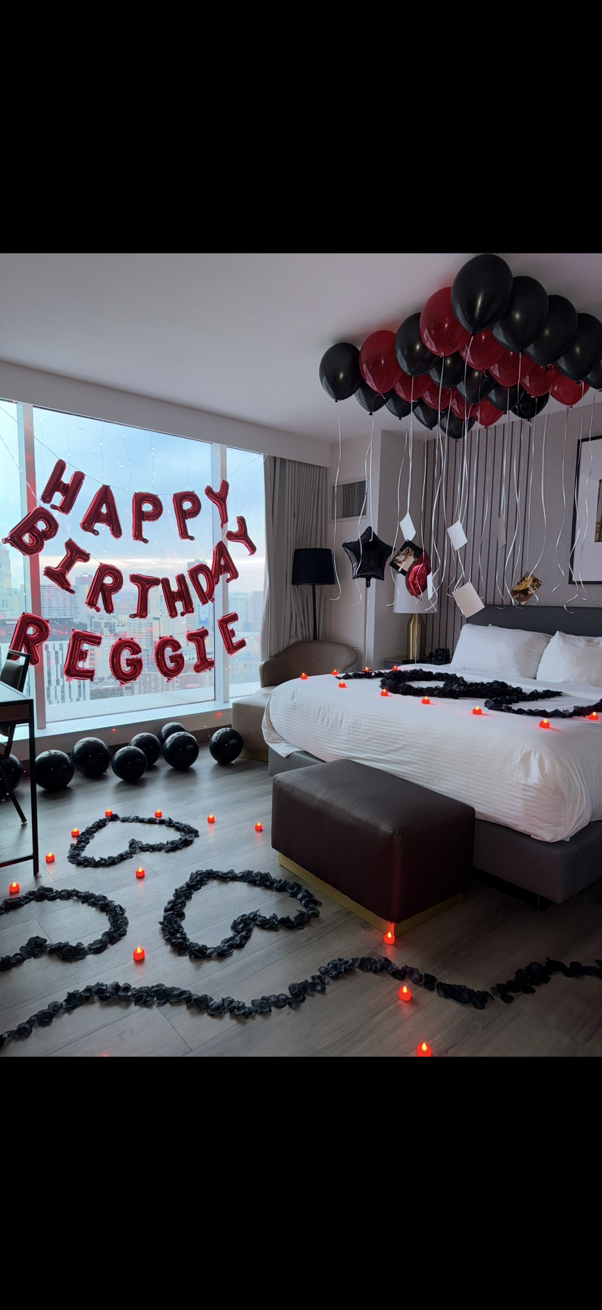 Hotel room decorated for a birthday celebration with red and black balloons, candles, and a "Happy Birthday Reggie" balloon banner, city view through large window.