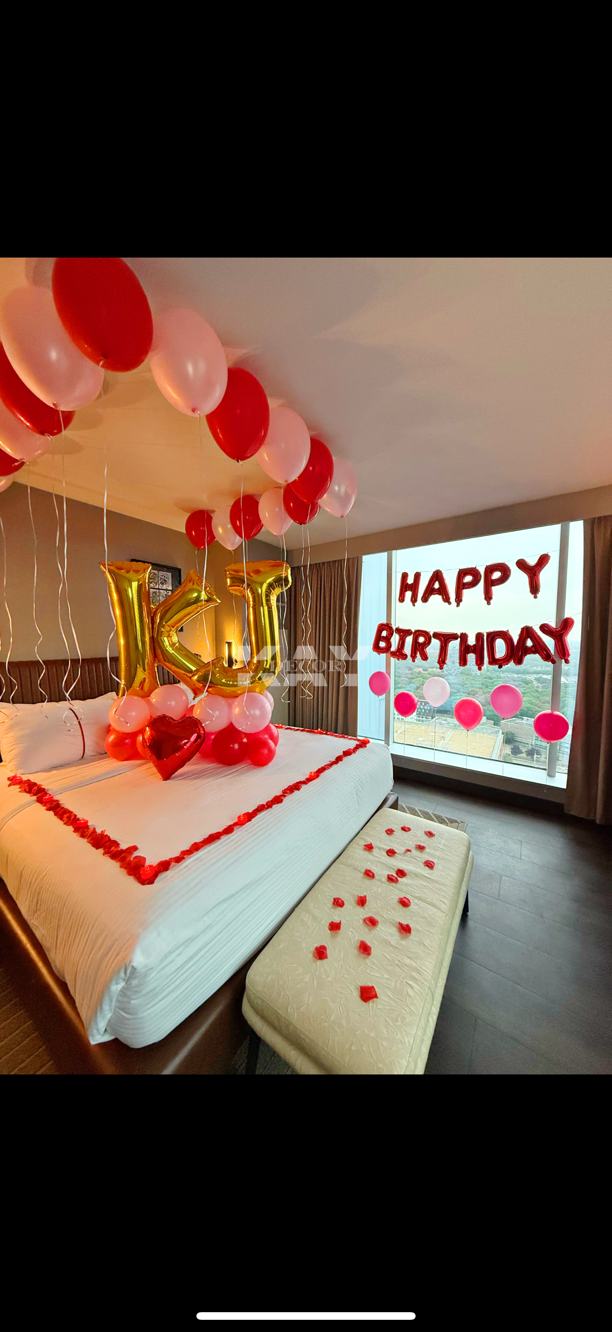 Decorated hotel room with balloons, 'HAPPY BIRTHDAY' banner, balloon heart, rose petals on the bed, and large gold balloons forming the initials 'KJ'.