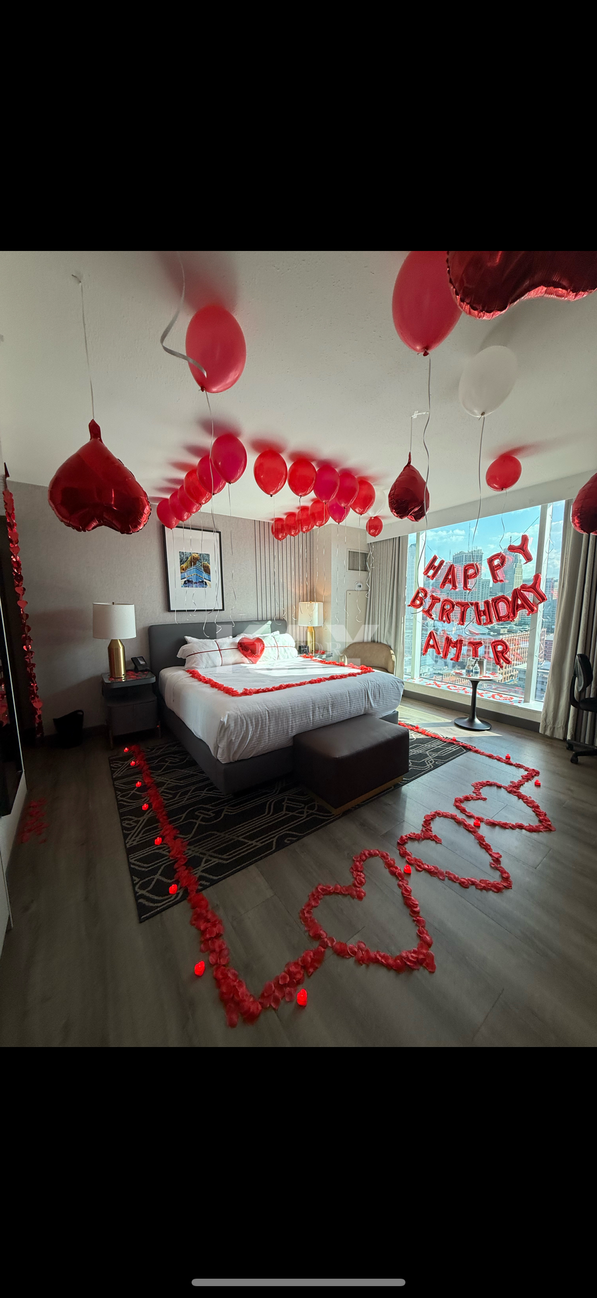 Hotel room decorated for a birthday with red and pink balloons, rose petals, a "Happy Birthday" banner, and heart-shaped accents.