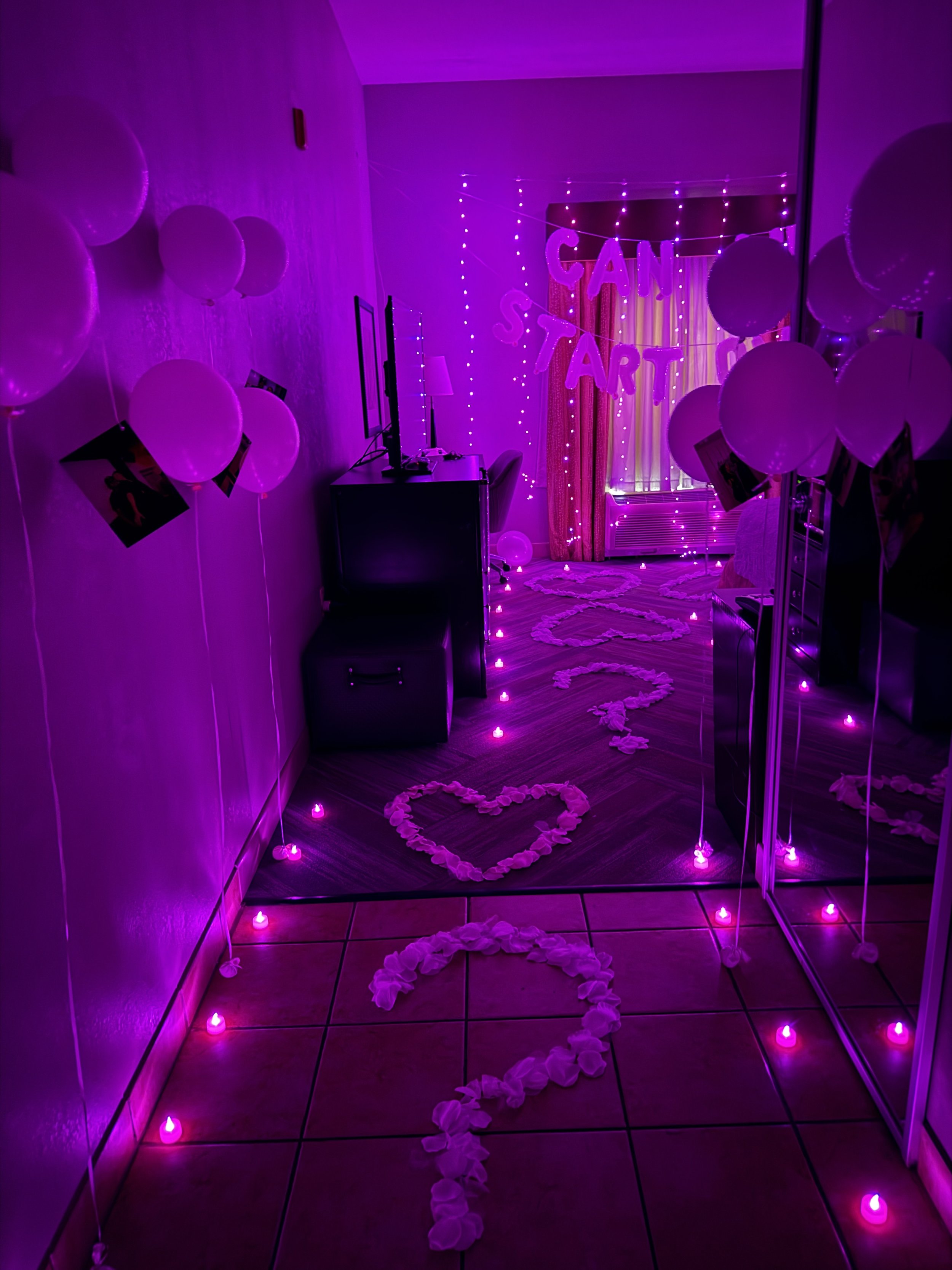 A room decorated with pink and purple lights, balloons, and heart-shaped flower arrangements on the floor for a celebration, with a large “GANT START” sign and a curtain in the background.