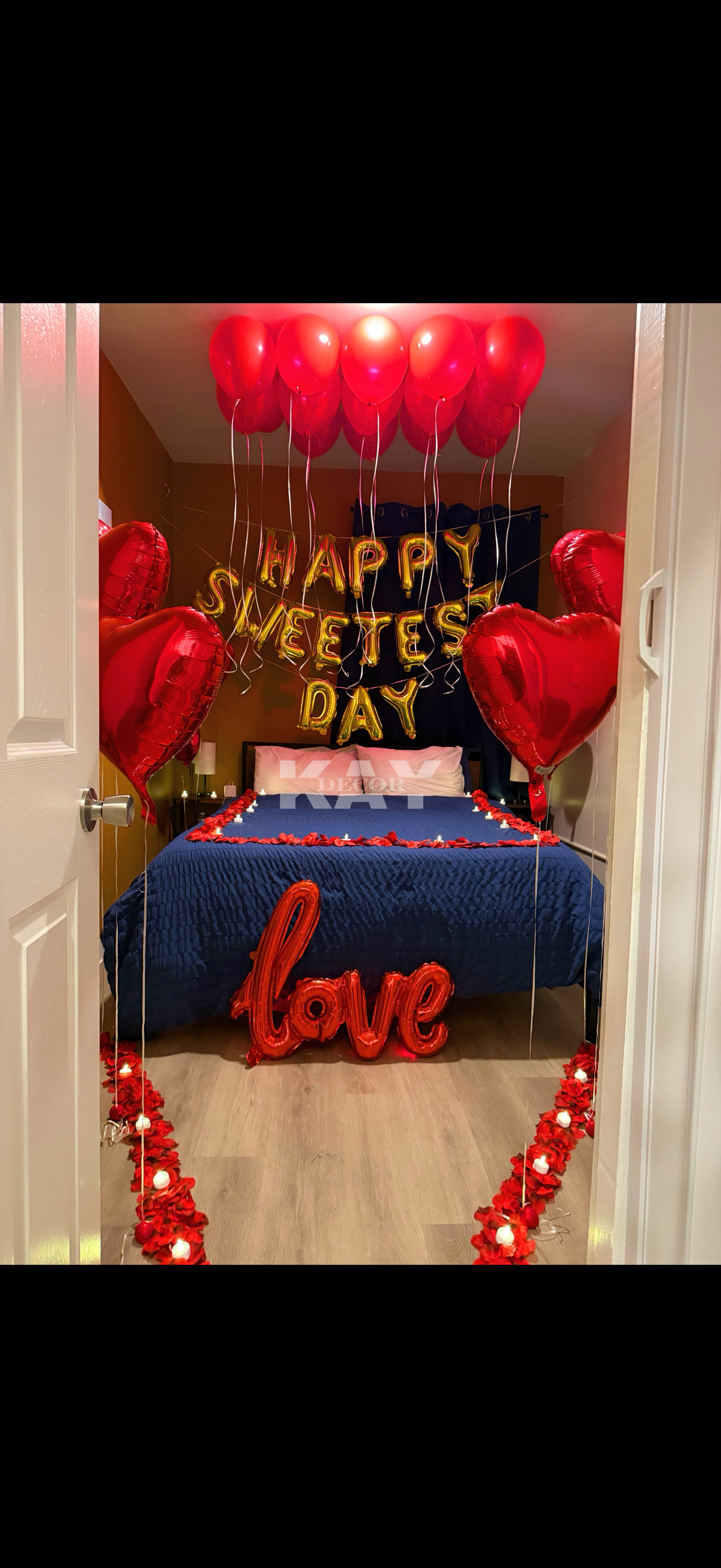 Valentine's Day decorated bedroom with red heart-shaped balloons, gold letter balloons reading "Happy Sweetest Day," and a red "love" balloon on the bed. Red flower petals and candles outline the floor leading to the bed.