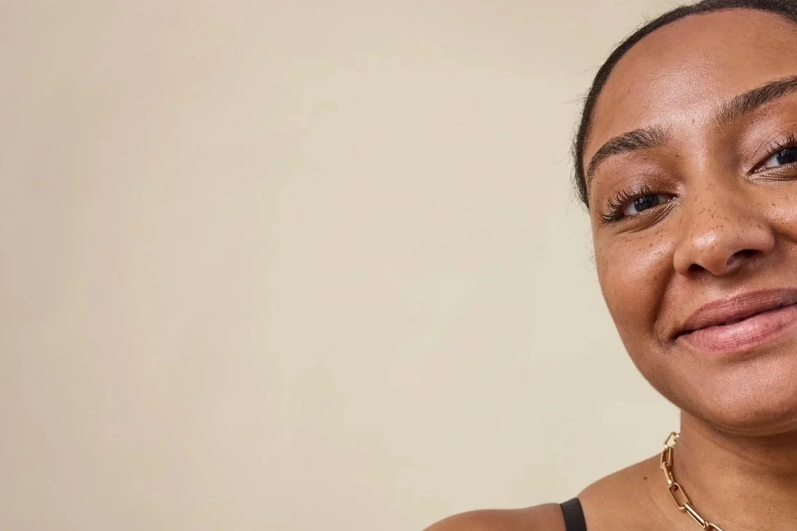 Close-up of a smiling woman with brown skin, dark hair, and a gold chain necklace, against a plain beige background.