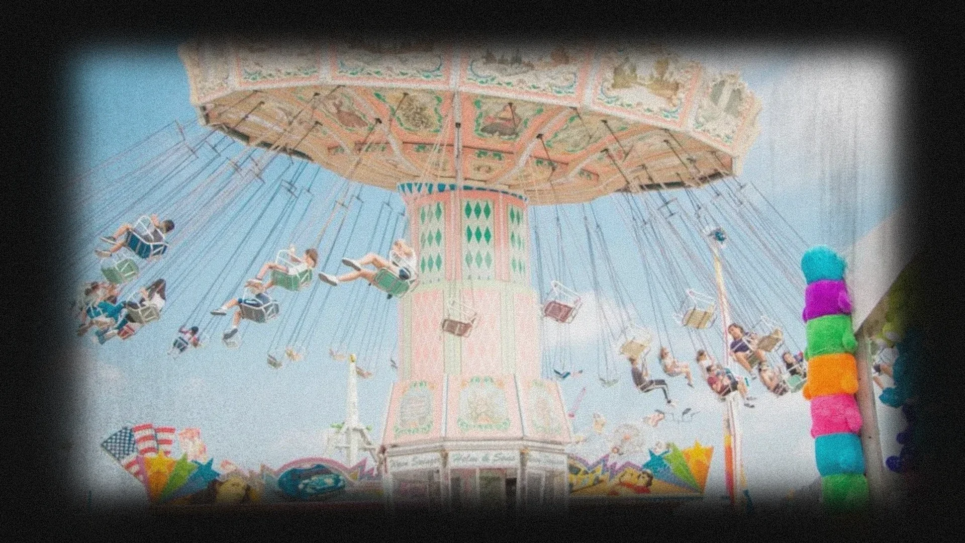 Swing ride at a fair with people enjoying the ride, surrounded by colorful decorations and a stall with stuffed animals.