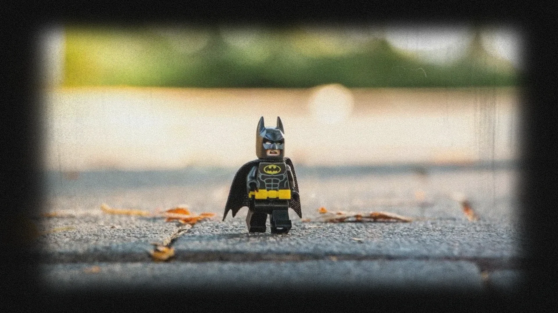 Lego Batman mini-figure standing on sidewalk with autumn leaves, blurred greenery background.