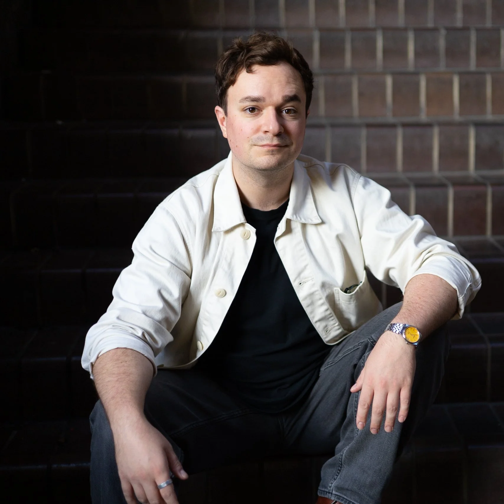 A young man sitting on dark brick stairs, wearing a cream-colored jacket, black shirt, jeans, and a yellow watch.
