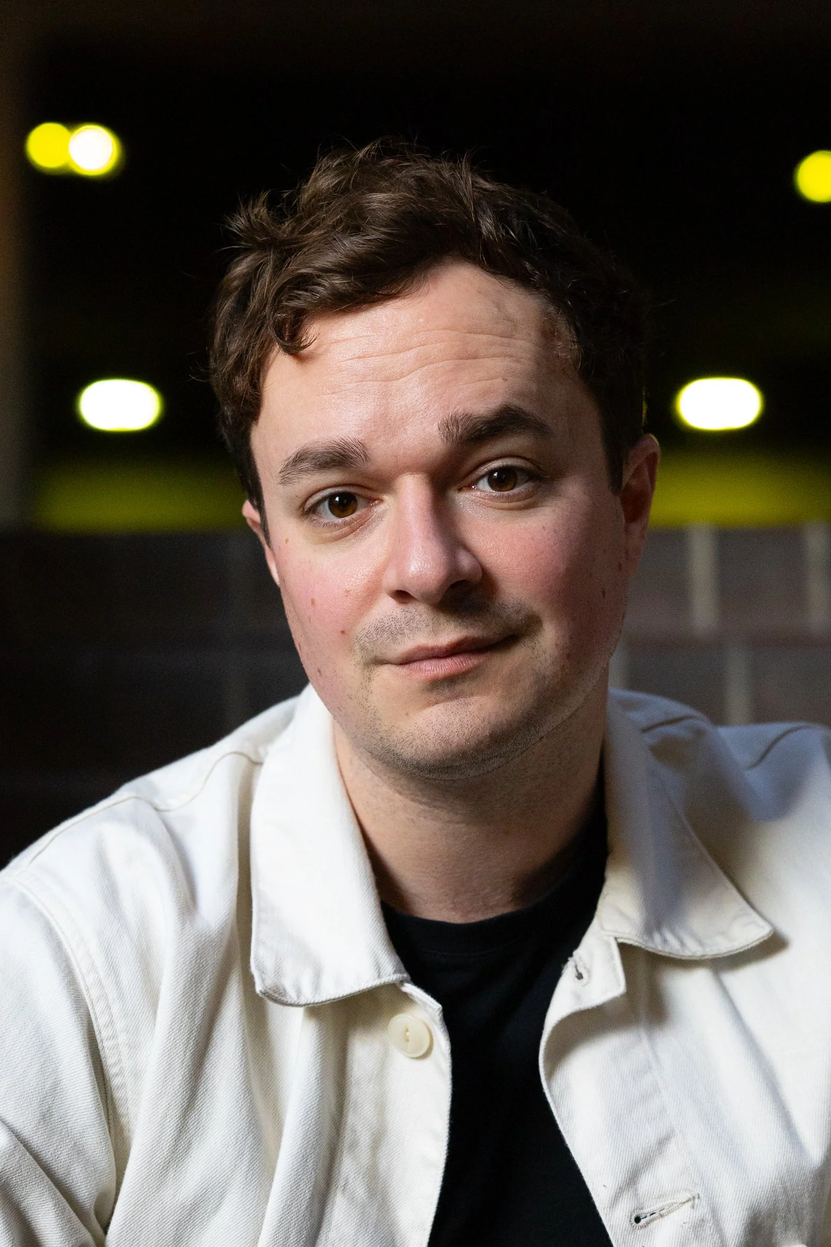A young man with brown hair and a slight smile, wearing a white jacket over a black shirt, sitting indoors with blurred yellow lights in the background.