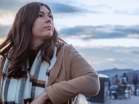 A young woman with long brown hair, wearing a brown coat and a striped scarf, looking off into the distance outdoors with mountains and a cloudy sky in the background.