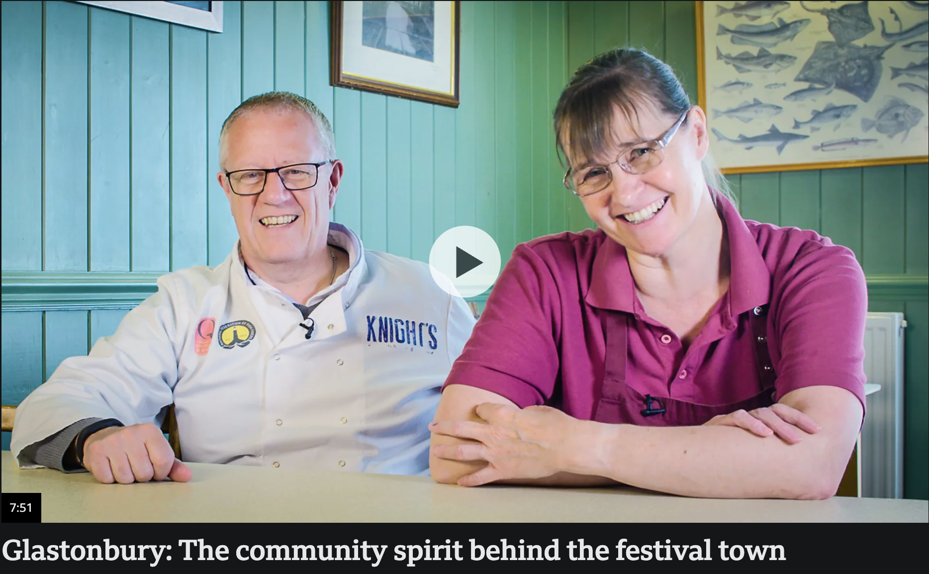 A man and a woman sitting at a table, smiling and looking at the camera. The man is wearing glasses and a chef's coat with patches, and the woman is wearing glasses and a purple shirt. They are in a room with green paneling on the wall, and there are framed pictures of fish and a landscape behind them.