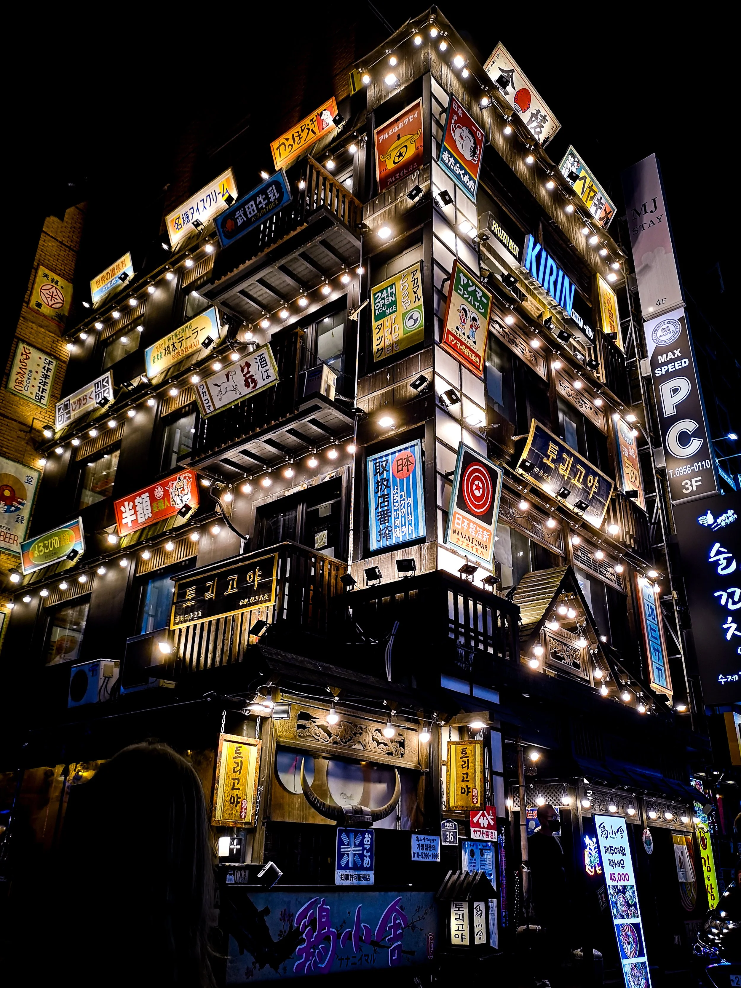 Night shot of a multi-story building with numerous illuminated signs in Japanese and English, storefronts, and balconies, with bright lights and a dark sky background.