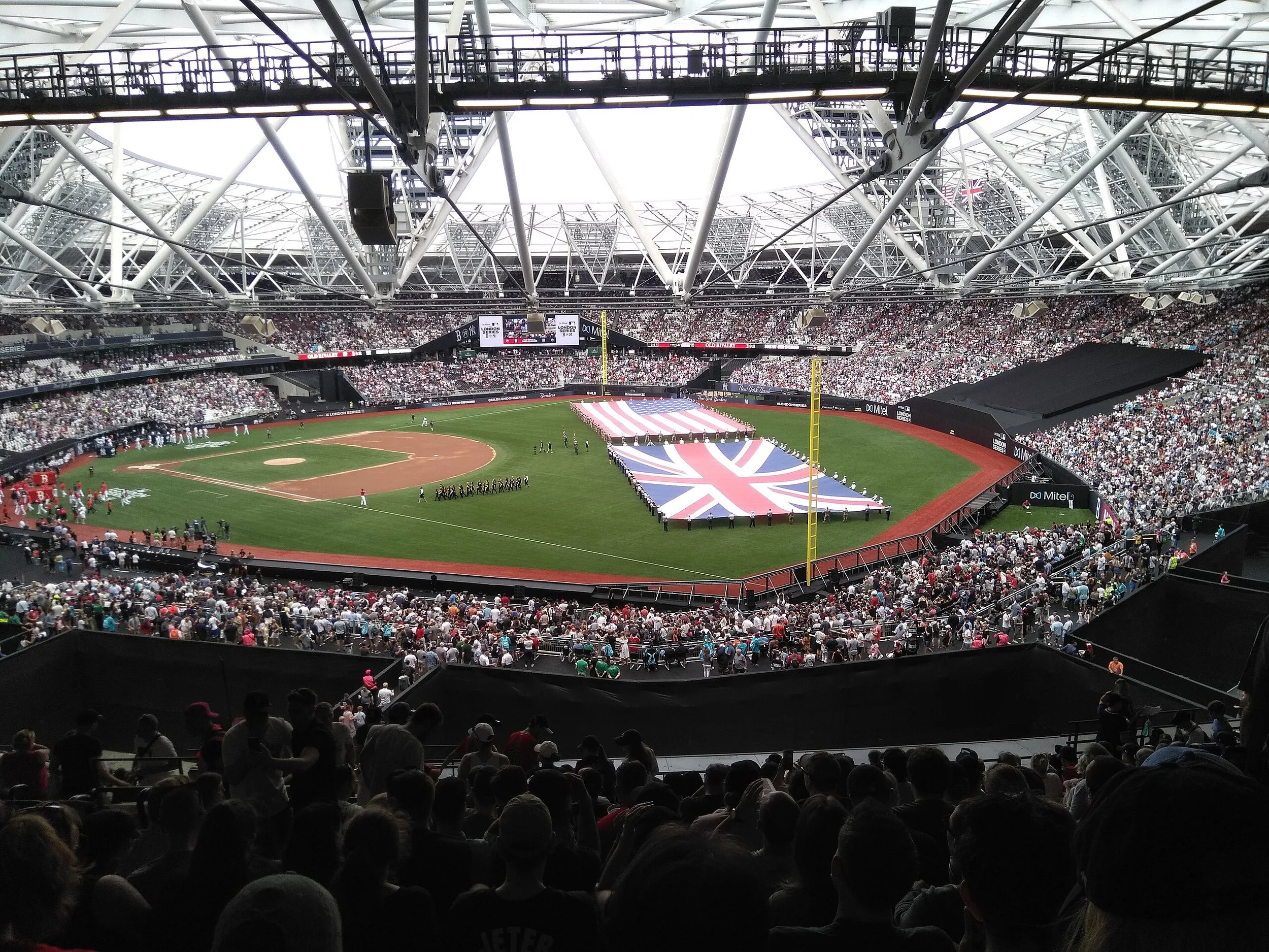 A MLB baseball stadium filled with spectators, with a large American flag being displayed on the field for a pre-game ceremony.