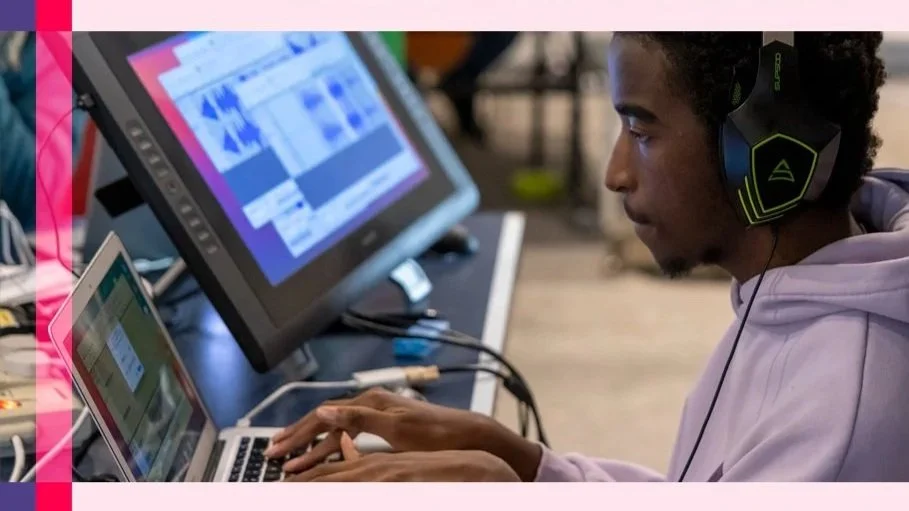 A person wearing a hoodie and gaming headset working on a laptop and desktop computer in a modern workspace.