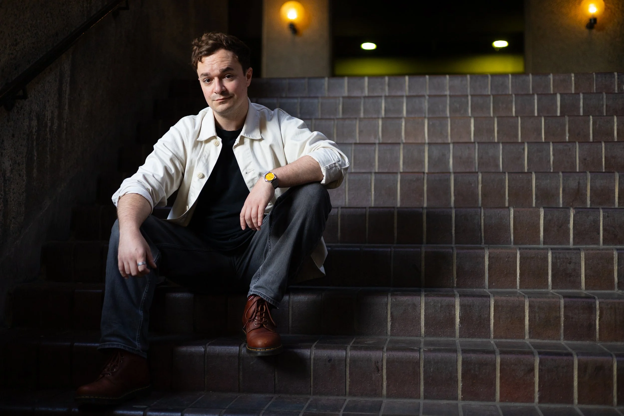 A young man sitting on tiled stairs in an indoor setting, wearing a white jacket, black shirt, gray jeans, and brown boots, with a watch on his left wrist, looking at the camera.
