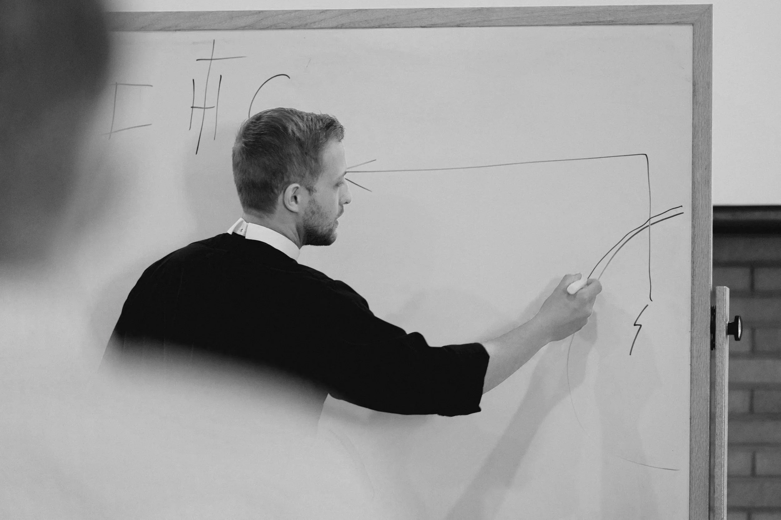 A pastor in a dark shirt writes on a whiteboard with a marker, drawing a line of life outside of Christ (IHC), while people watch.