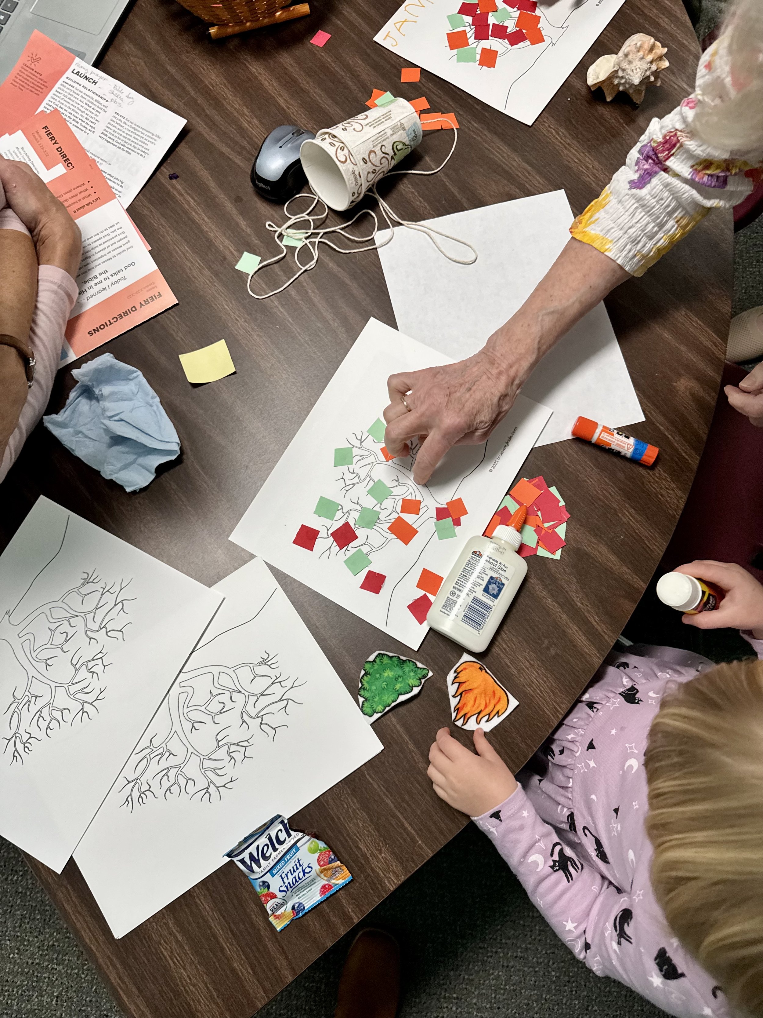 People engaging in a craft activity at a table, making a paper tree. The table has various craft supplies, including colored paper squares, glue sticks, markers, and printed tree templates. One person is placing colored paper on the tree template, while children and adults participate.