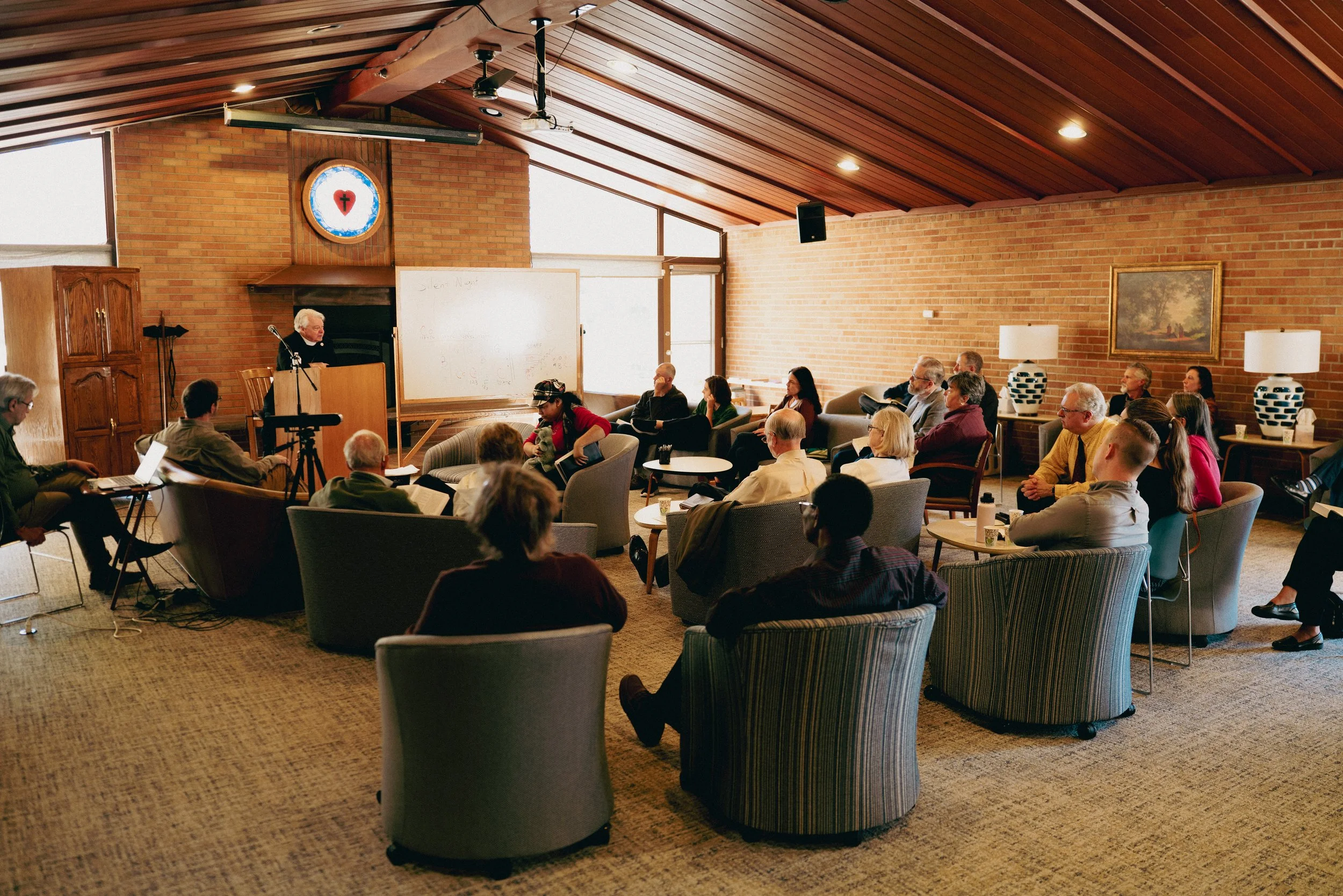 A group of people attending a bible study in our Fireside Room with brick walls and wooden ceiling, facing the pastor at a podium near a whiteboard.