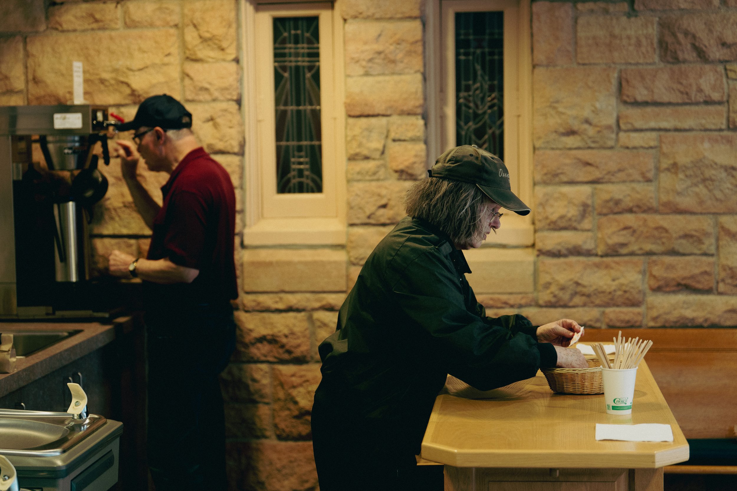 An woman wearing a black jacket and cap stands at a wooden counter setting up for coffee hour.