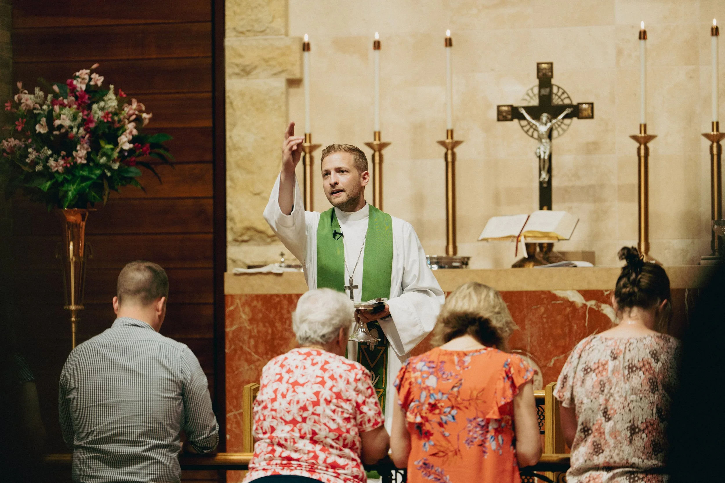 A pastor dressed in a white robe and green stole is blessing people with a cross hand gesture at the communion rail.