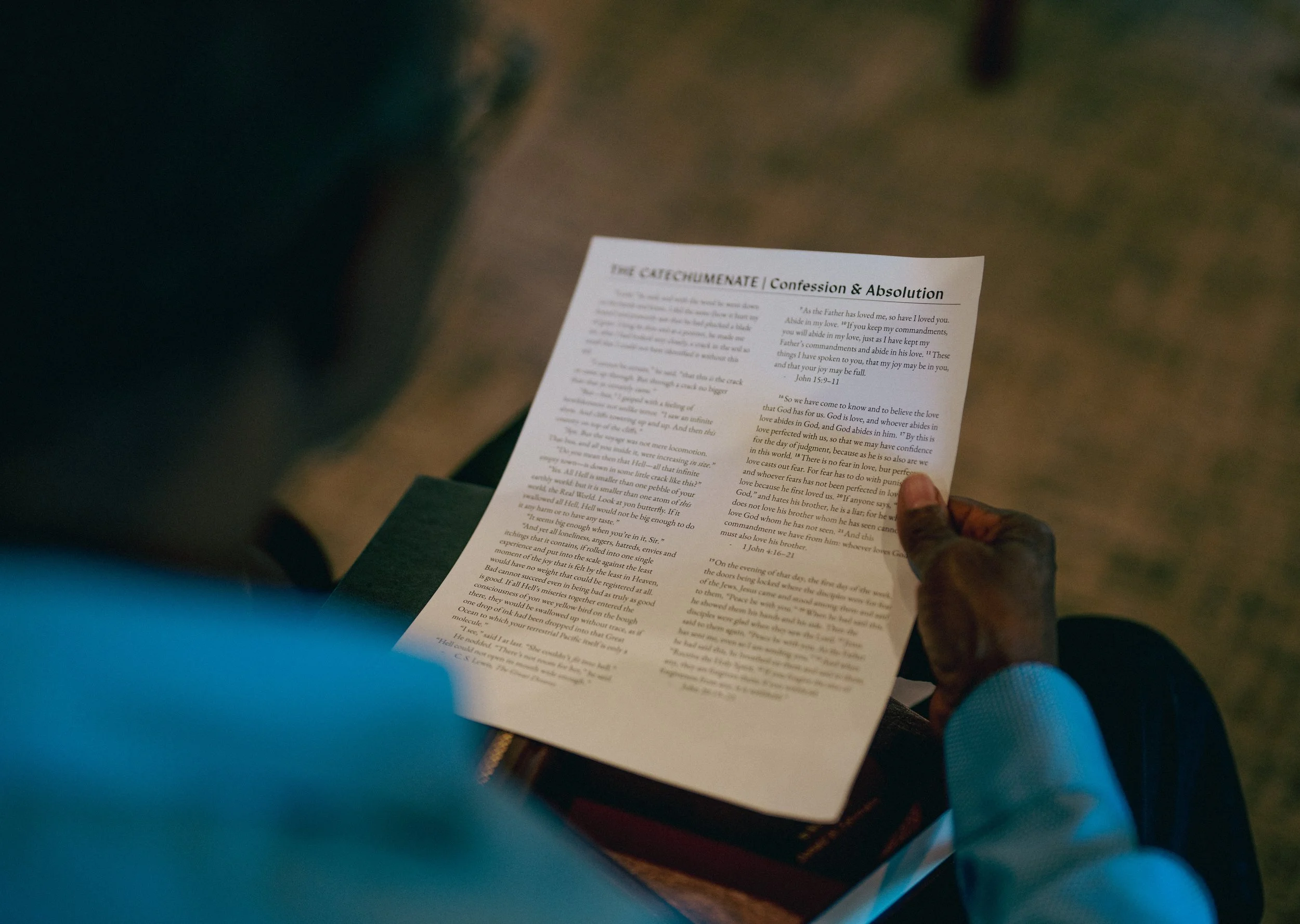 A person holding a printed religious or religious-themed document discussing 'The Catechumenate: Confession and Absolution'. The person is sitting down, wearing a light-colored long sleeve shirt, and the background shows a patterned carpeted floor.