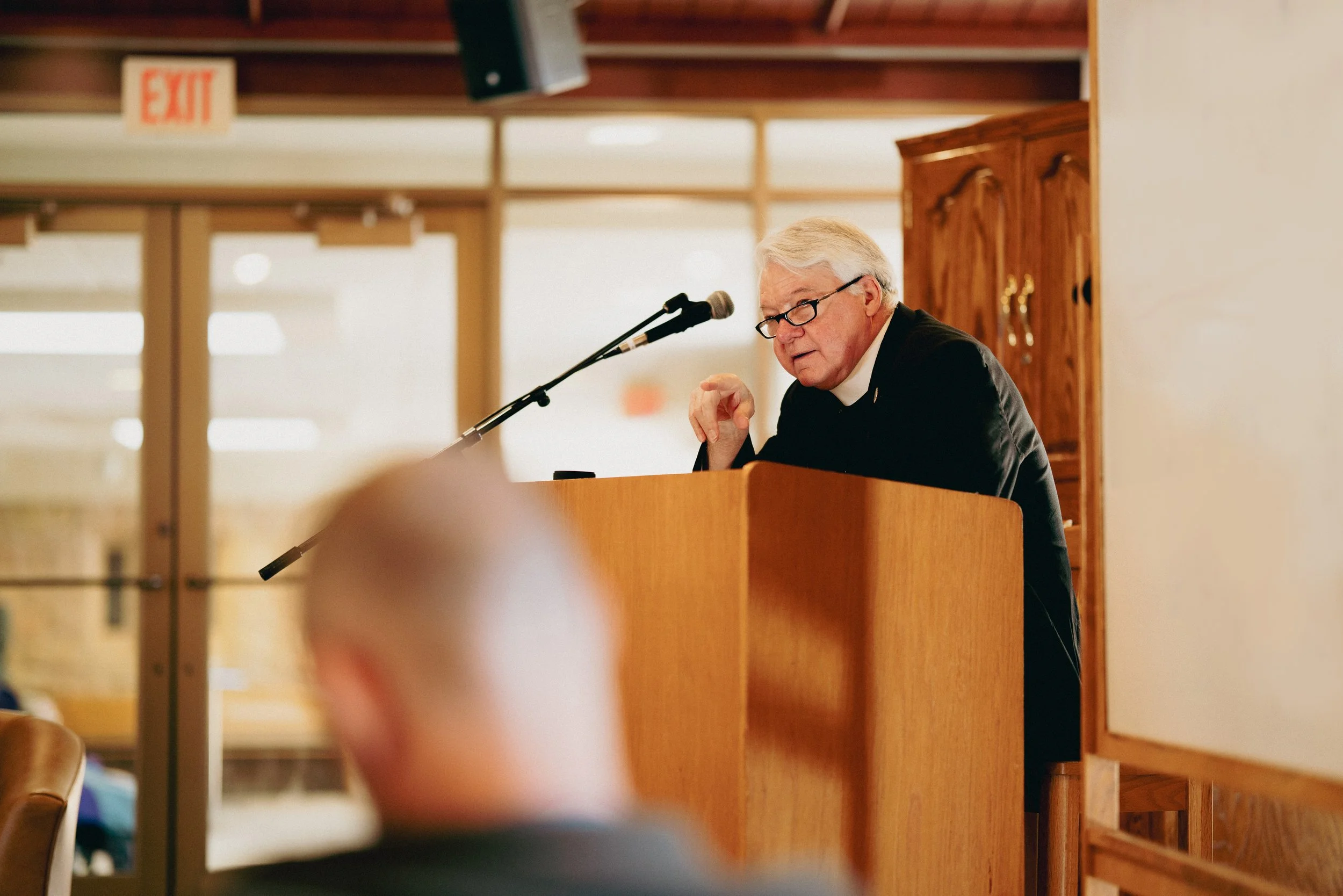 An elderly pastor with gray hair, glasses, and a black jacket speaking at a podium in a room with wooden cabinets and glass doors.