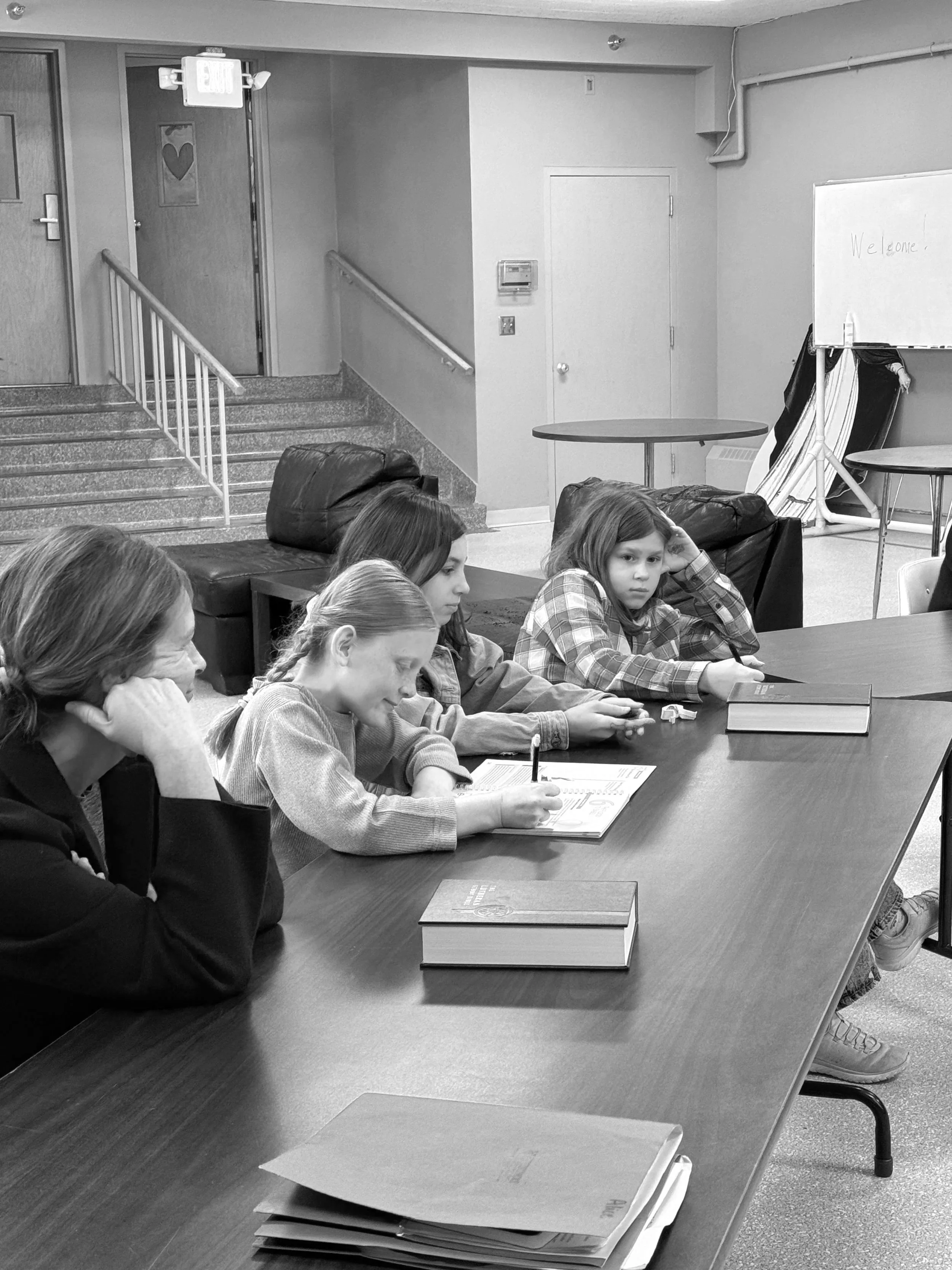 Four children and one adult sitting at a long table in a classroom or meeting room, with books and papers in front of them. Two of the children are writing or drawing, one child looks pensive, and of the adults, one is resting her chin on her hand, observing the scene.
