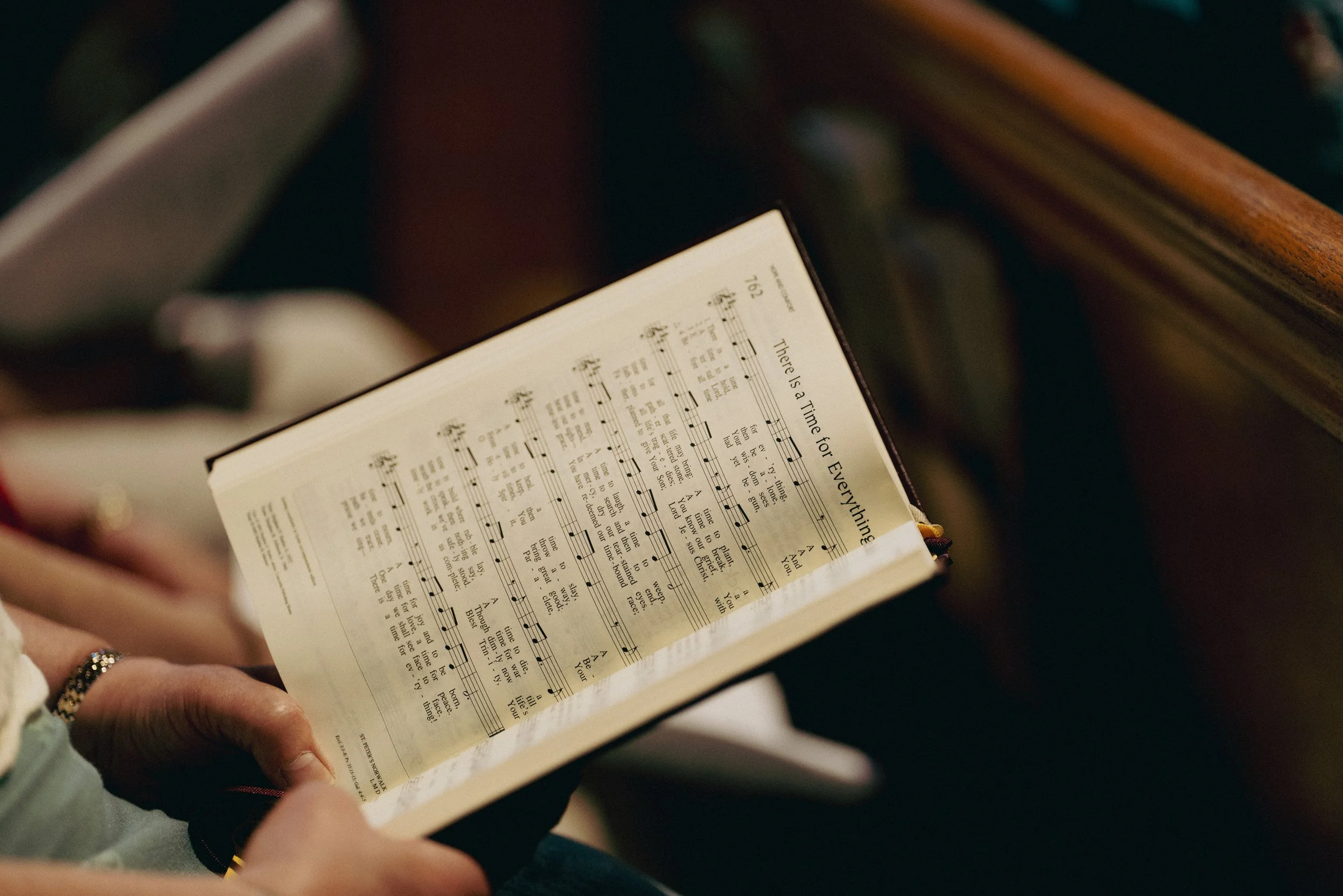 A person holding and reading a hymn book with sheet music and lyrics inside a church pew.