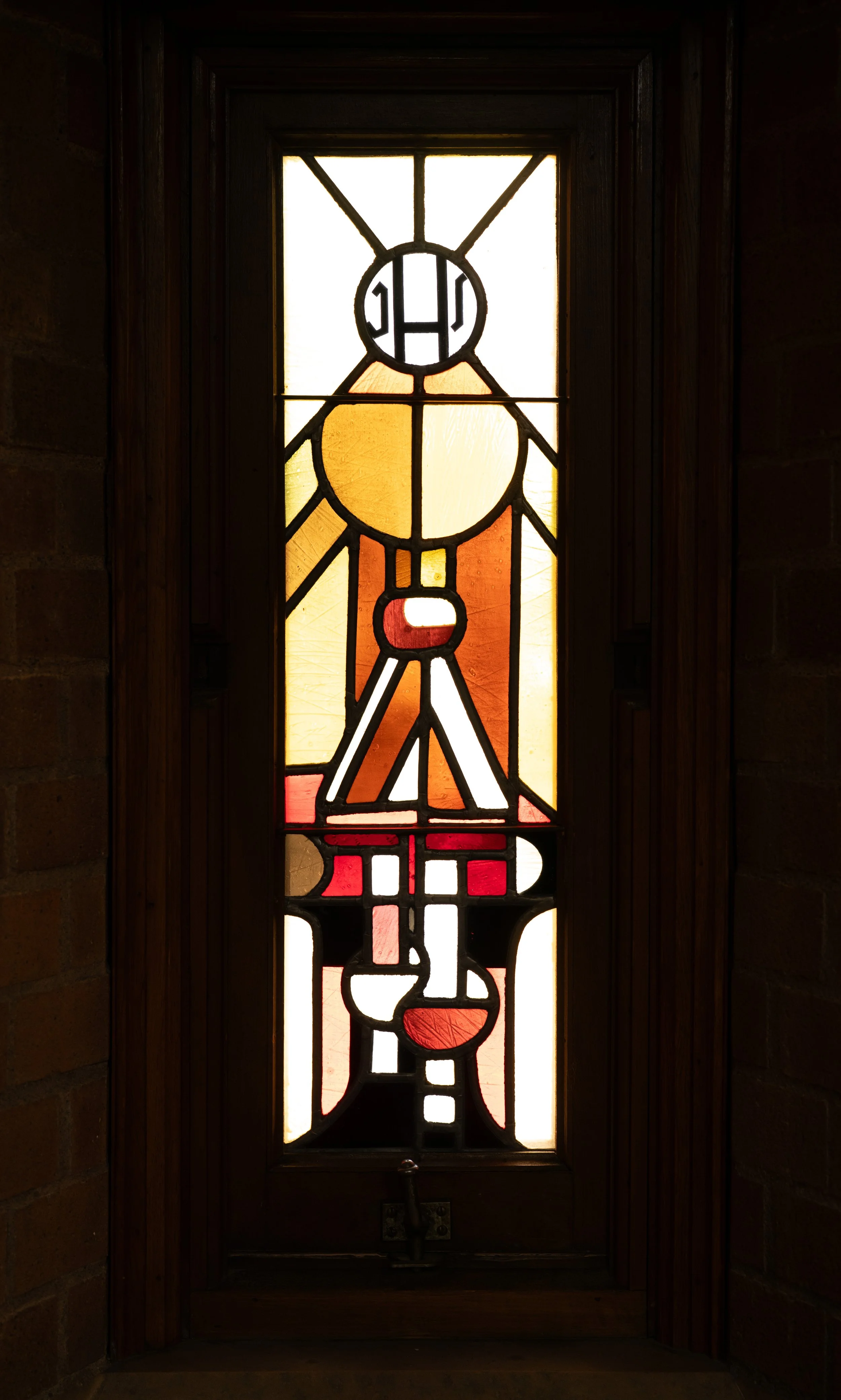 A stained glass window featuring abstract geometric shapes and patterns in yellow, orange, red, black, and white, framed by dark wood.