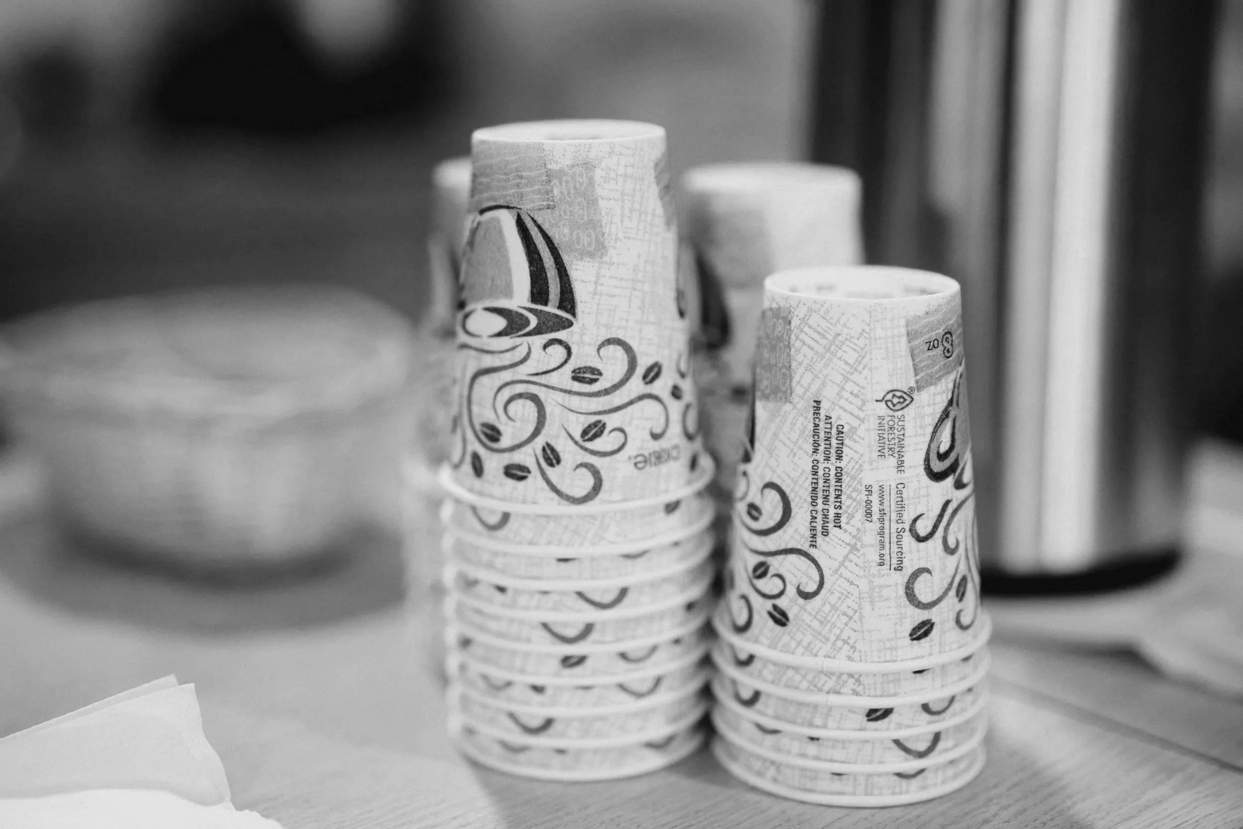 Stacks of coffee cups with decorative patterns on a table, with a blurry background of plates and a coffee pot.
