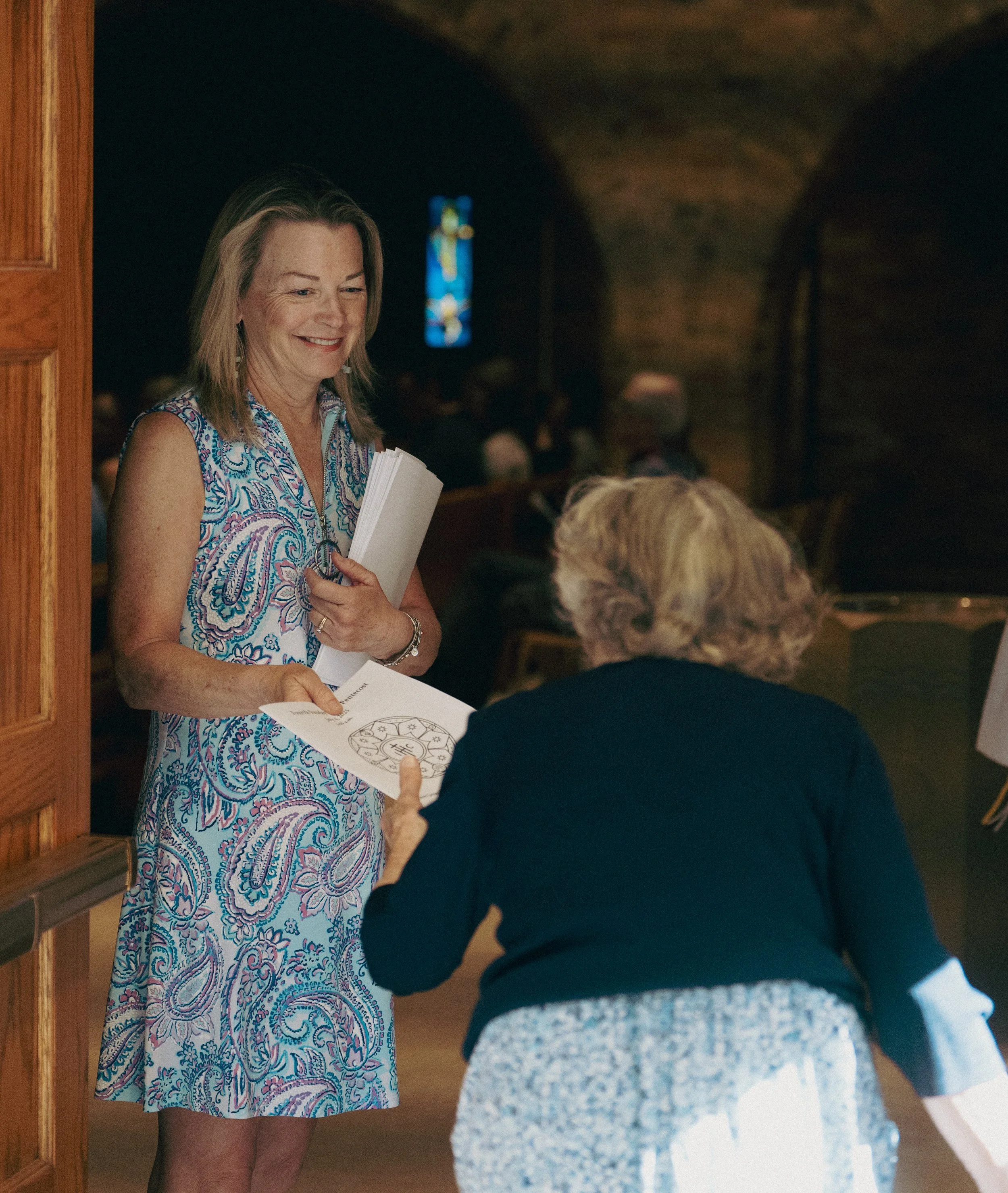 A woman standing and smiling while handing a service bulletin to an elderly woman in church with stained glass windows in the background.
