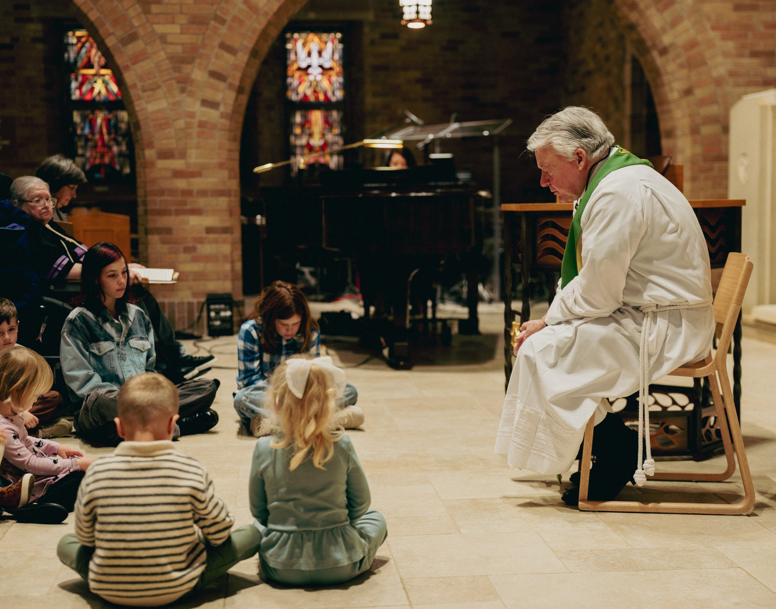 A pastor sitting on a chair with a green stole around his neck, sitting in front of children seated on the floor and adults seated on pews inside a church with stained glass windows.