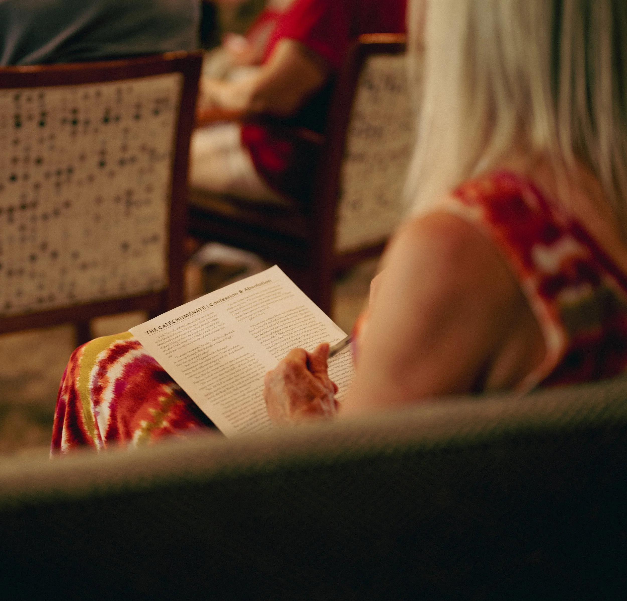 An older woman with blonde hair reading a pamphlet titled 'The Catechumenate: Confession and Absolution' in a room with people sitting in chairs.