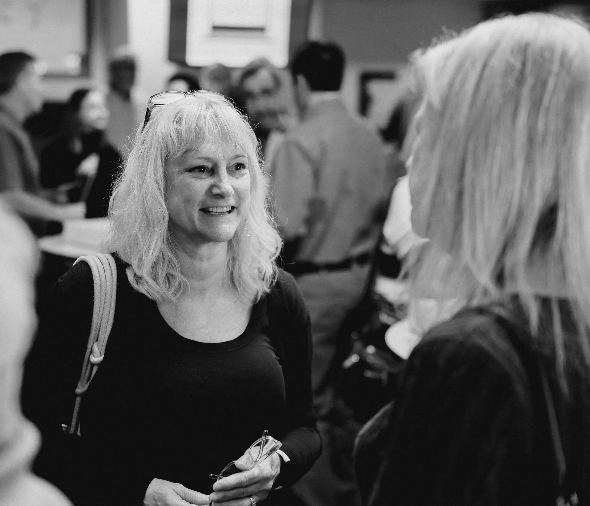 Black and white photo of a woman with blonde hair in a black top smiling and talking to another woman with long blonde hair in a black top, surrounded by a group of people in a room.