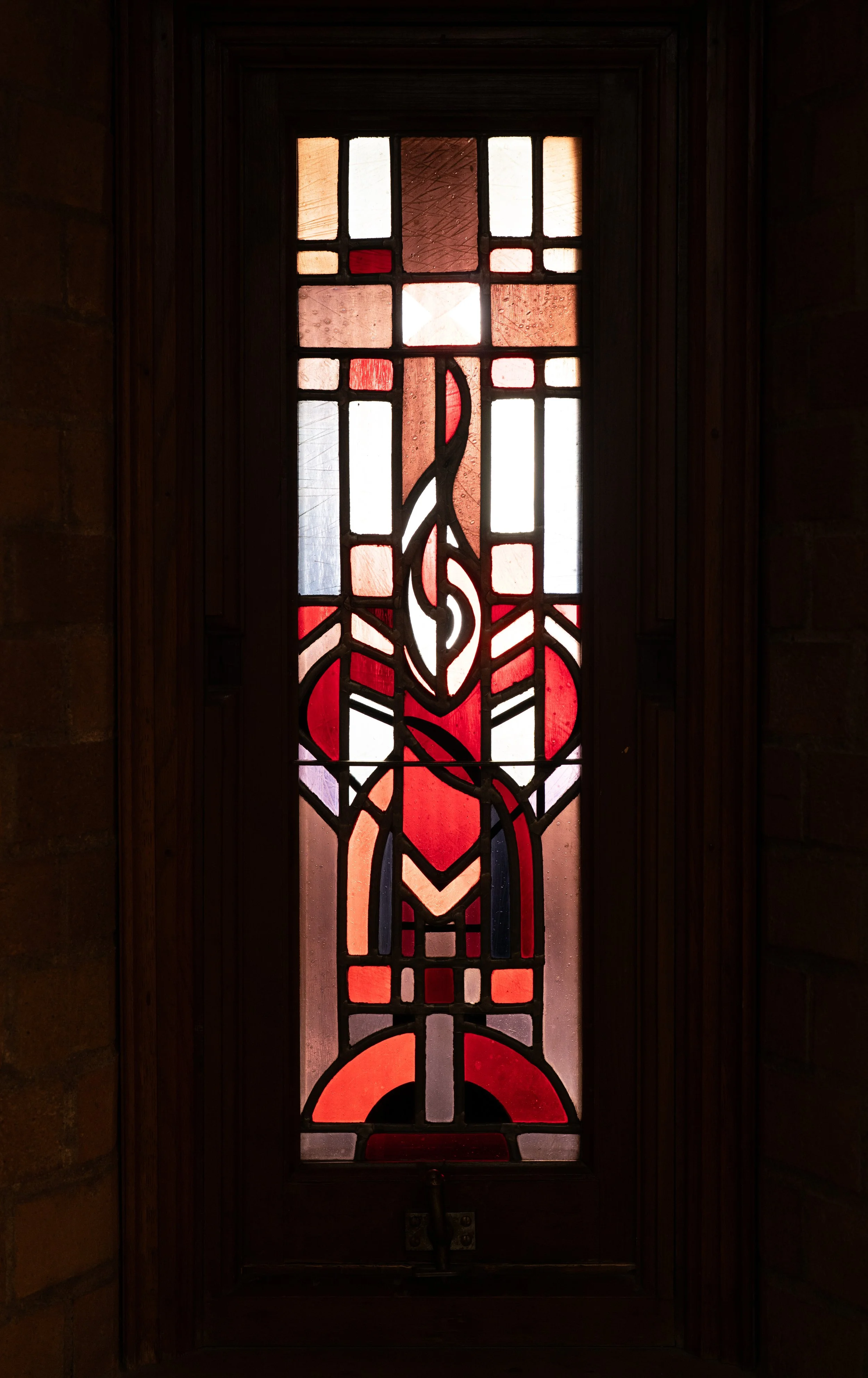 Stained glass window with abstract red, orange, purple, and white geometric patterns, framed by dark wood, with light filtering through.