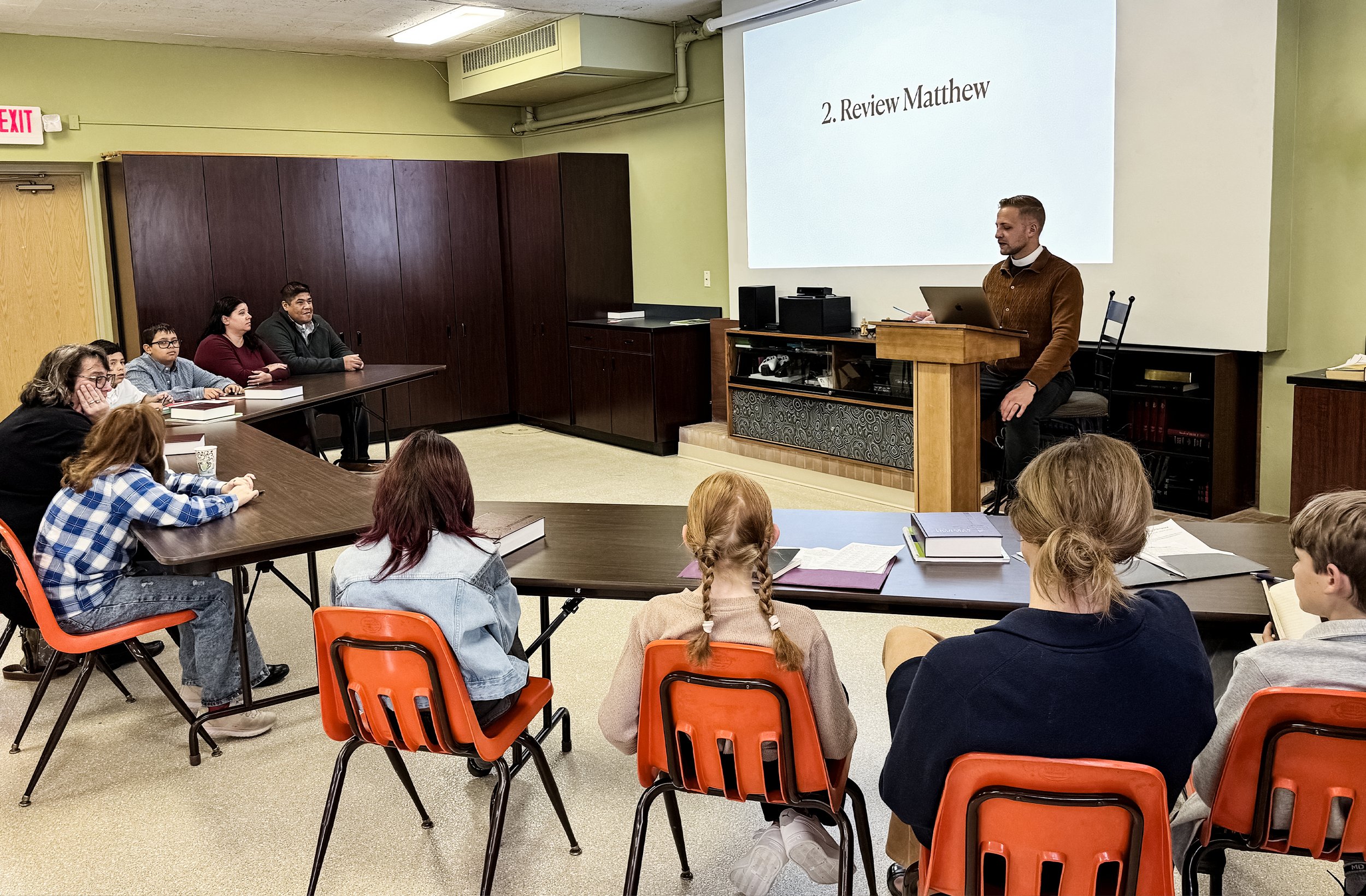 A pastor, teaching at a podium in front of a large screen with the words 'Review Matthew' displayed, addressing a group of students seated at tables in a classroom or meeting room.