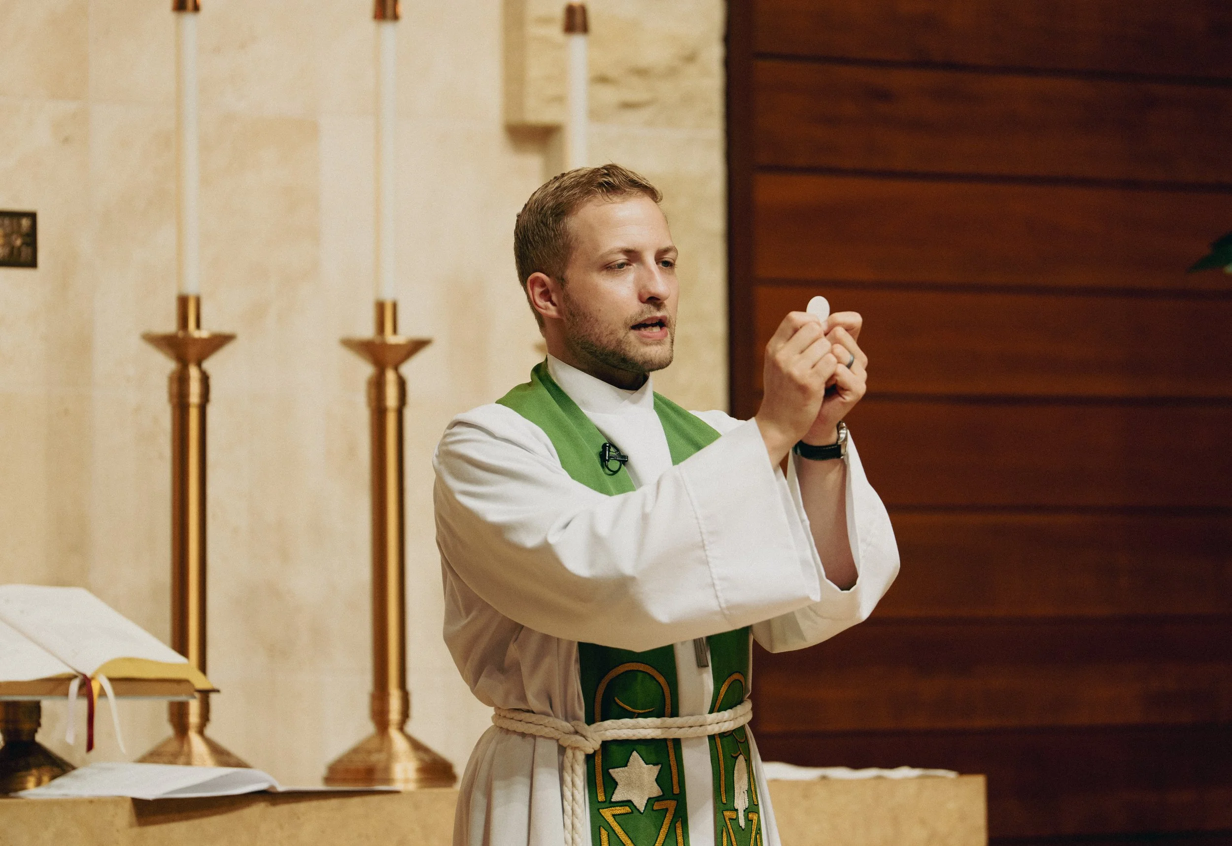 A pastor in white robes with green stole holding up the Eucharist during a church service, standing before altar with candles and open altar book.