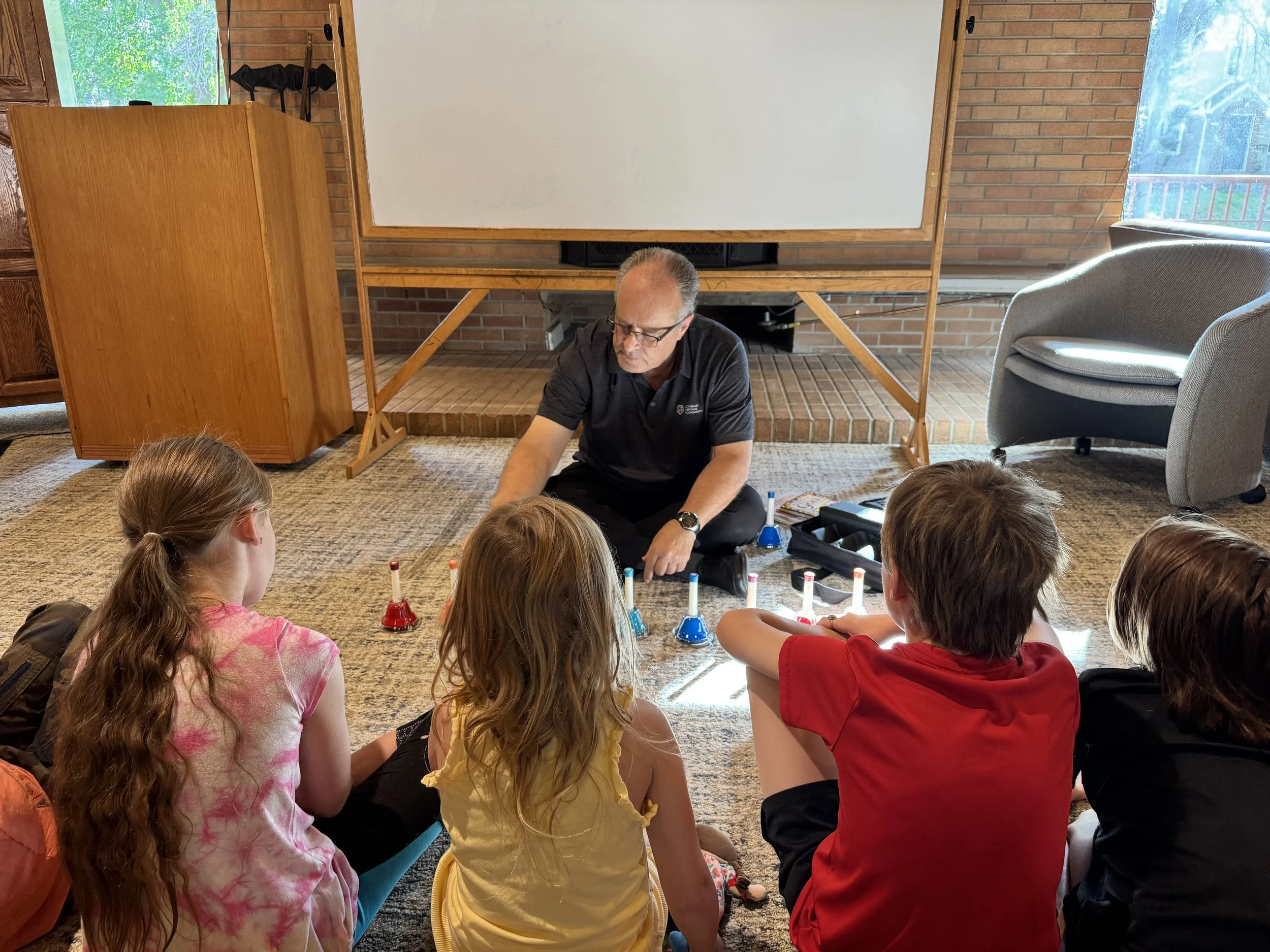 A man teaching a group of children about hand bells on a carpeted floor in a brightly lit room.