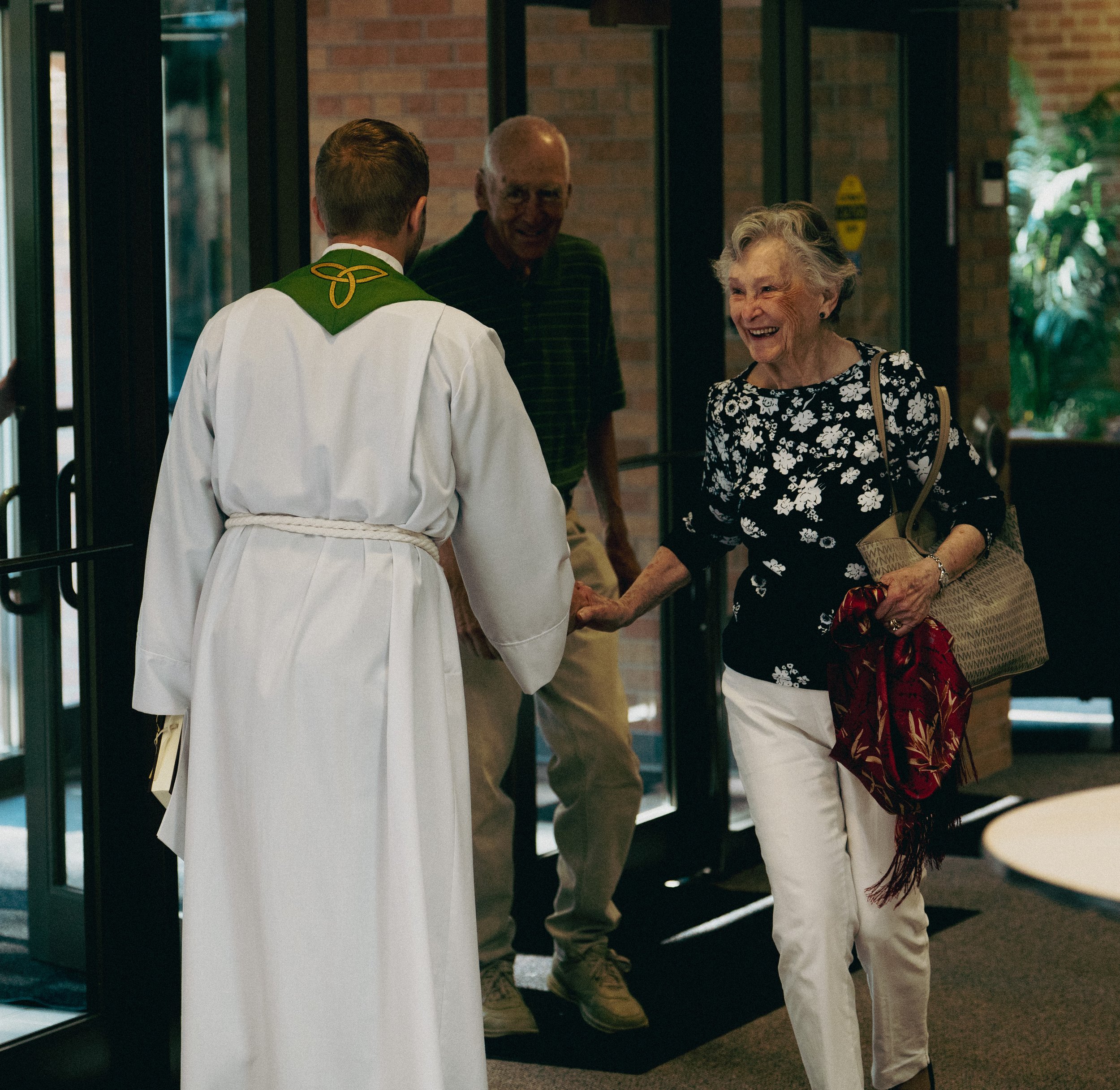An elderly woman in a black floral top and white pants greets the pastor in a white robe with green collar, holding his hand and smiling, while another man stands nearby at the entrance of a building.