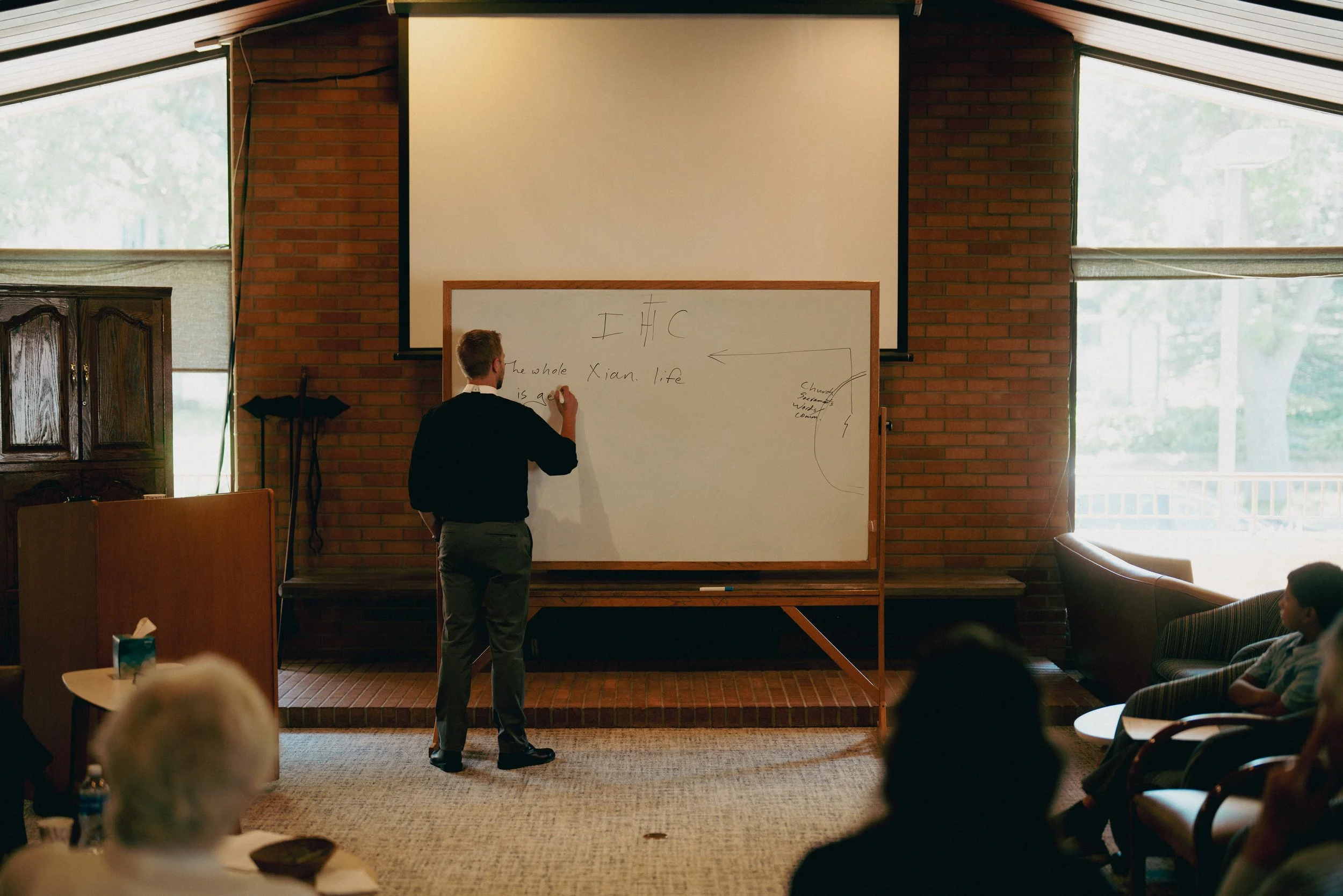 A pastor in a black clerical dress, writes on a whiteboard in front of a brick wall, during a bible study. Several people sit and watch, with large windows on both sides of the room letting in natural light.