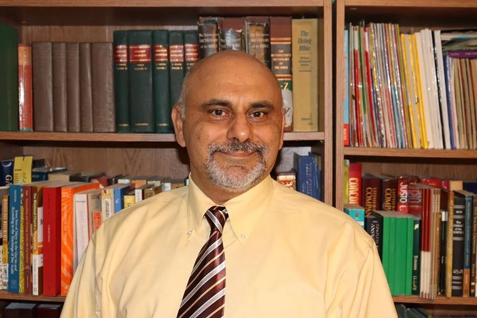 A man with a beard and mustache in a light yellow dress shirt and striped tie, standing in front of a wooden bookshelf filled with books.