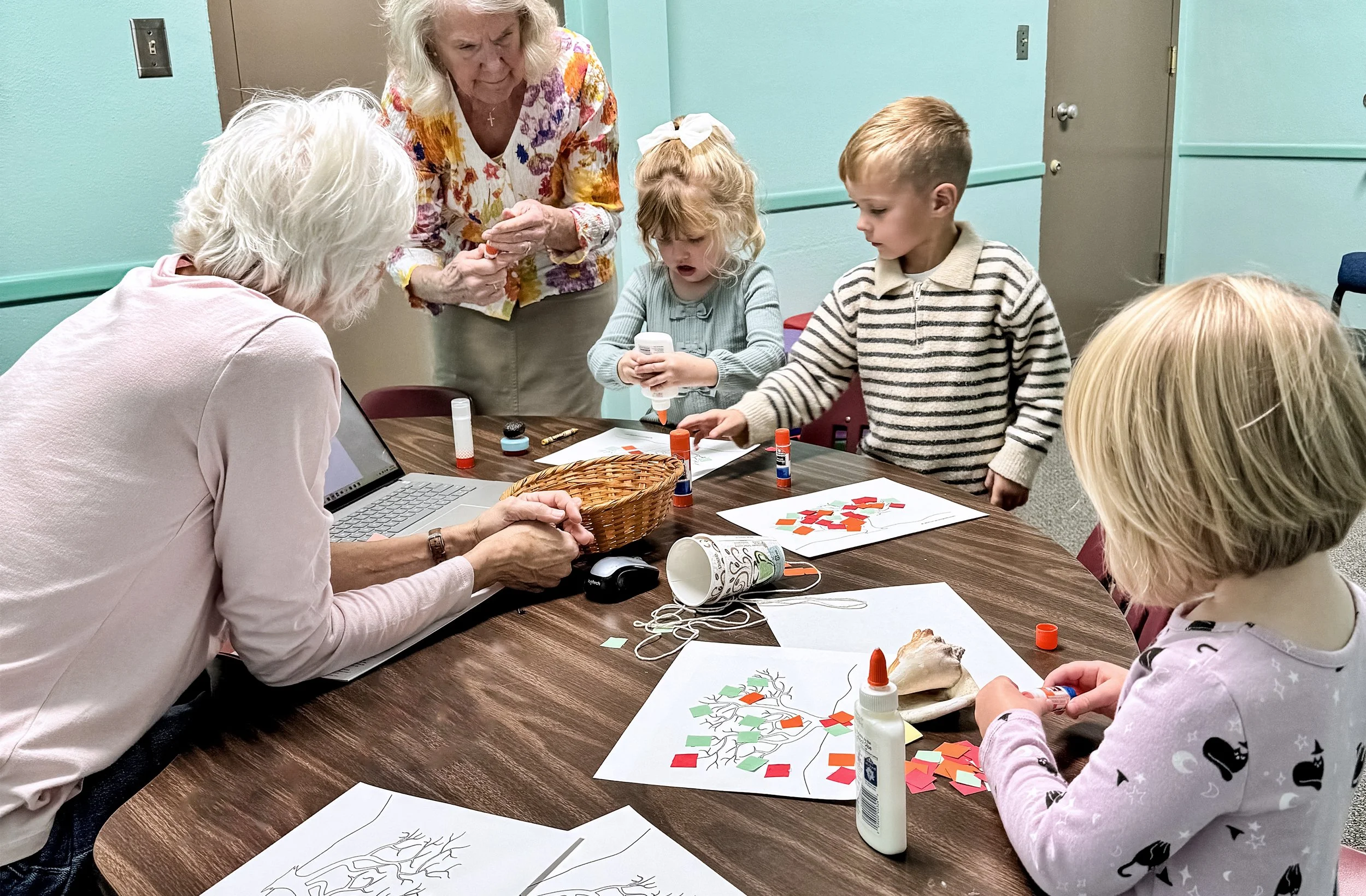 A group of children and two elderly women sitting around a table, engaging in arts and crafts activities including glue, paper, and leaves, in a room with mint green walls.