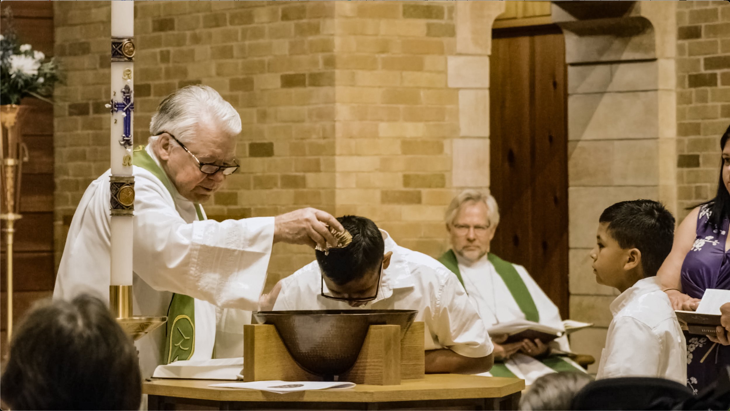 An elderly pastor with glasses pours water on the forehead of a boy during a baptism.