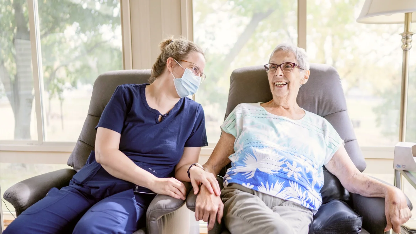 A healthcare worker, wearing blue scrubs and a face mask, holding hands and talking with an elderly woman sitting in a recliner chair near large windows with a view of trees outside. The elderly woman is smiling and wearing glasses and a patterned hospital gown.
