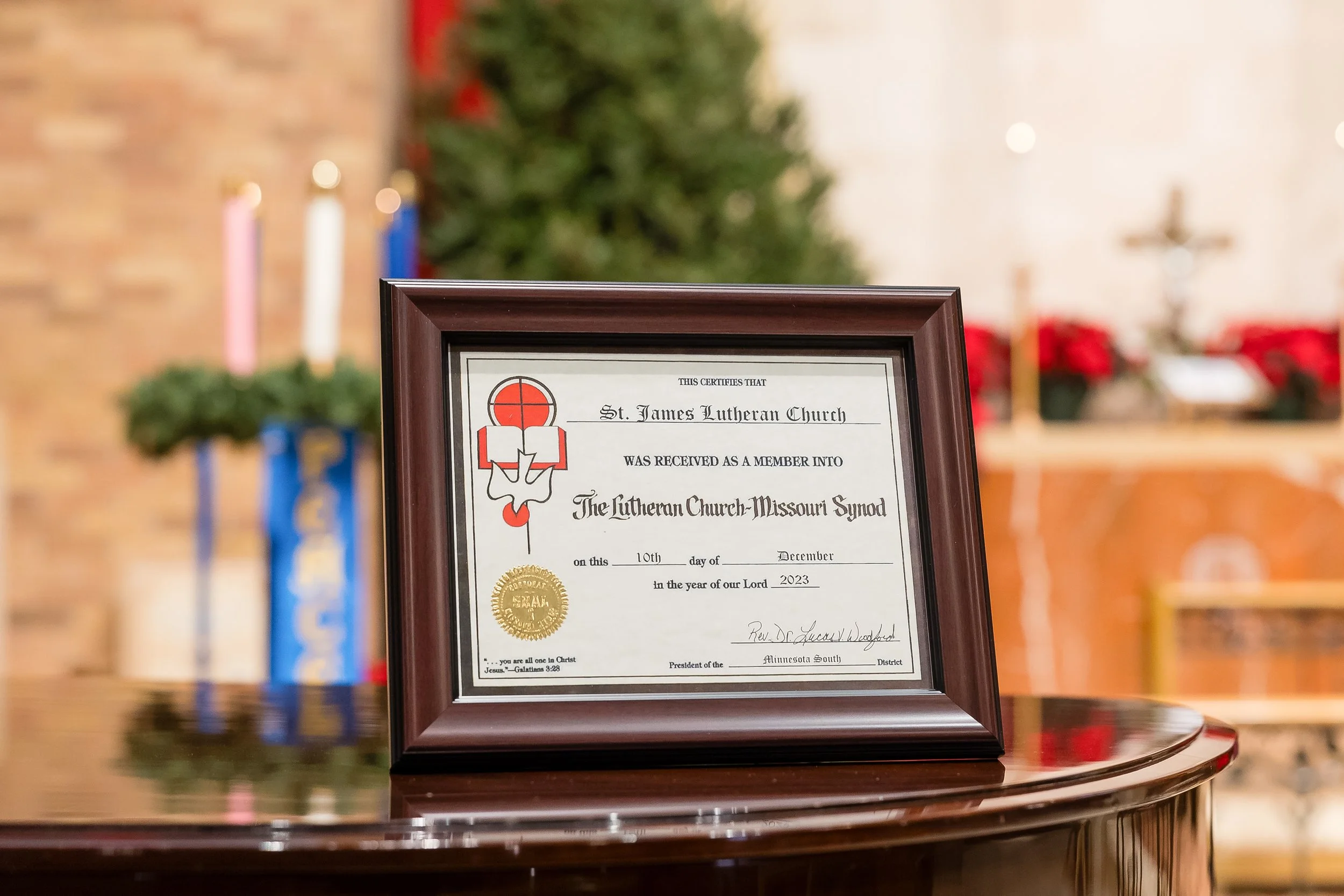 Framed certificate on a polished wooden table, with blurred Christmas tree and candles in background inside a church.