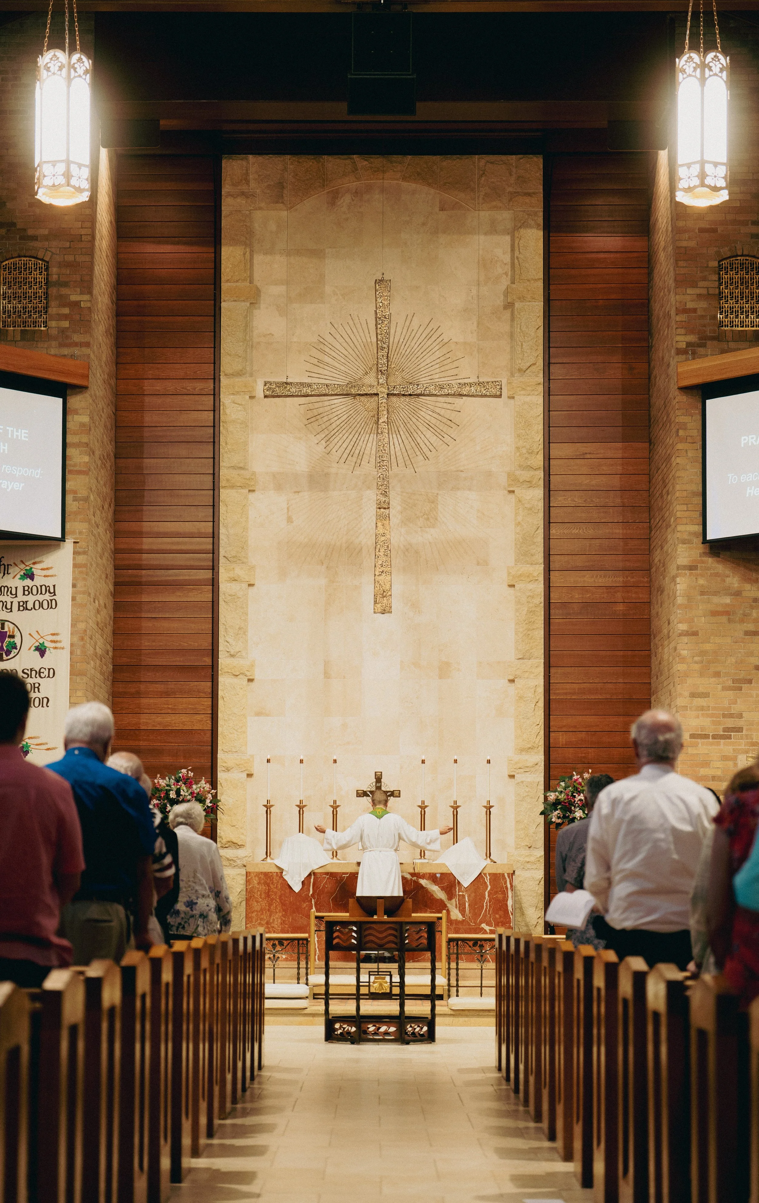People are gathered inside a church, standing in pews, facing the altar where the pastor is leading the service. The church has a large, decorated cross on the wall hanging above the altar.