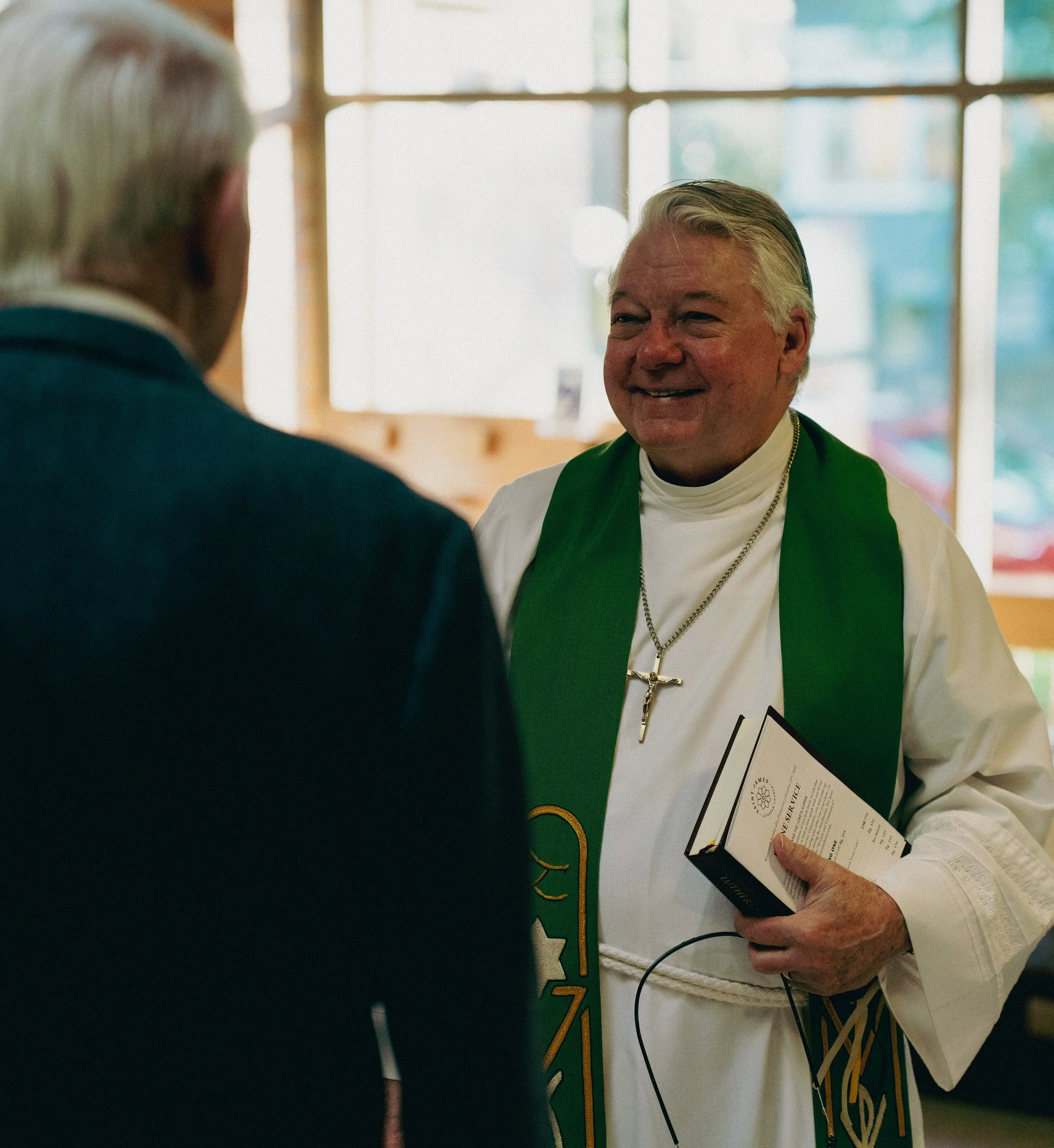 A pastor smiling and holding the LSB service book while talking to an older man with gray hair in the church narthex.