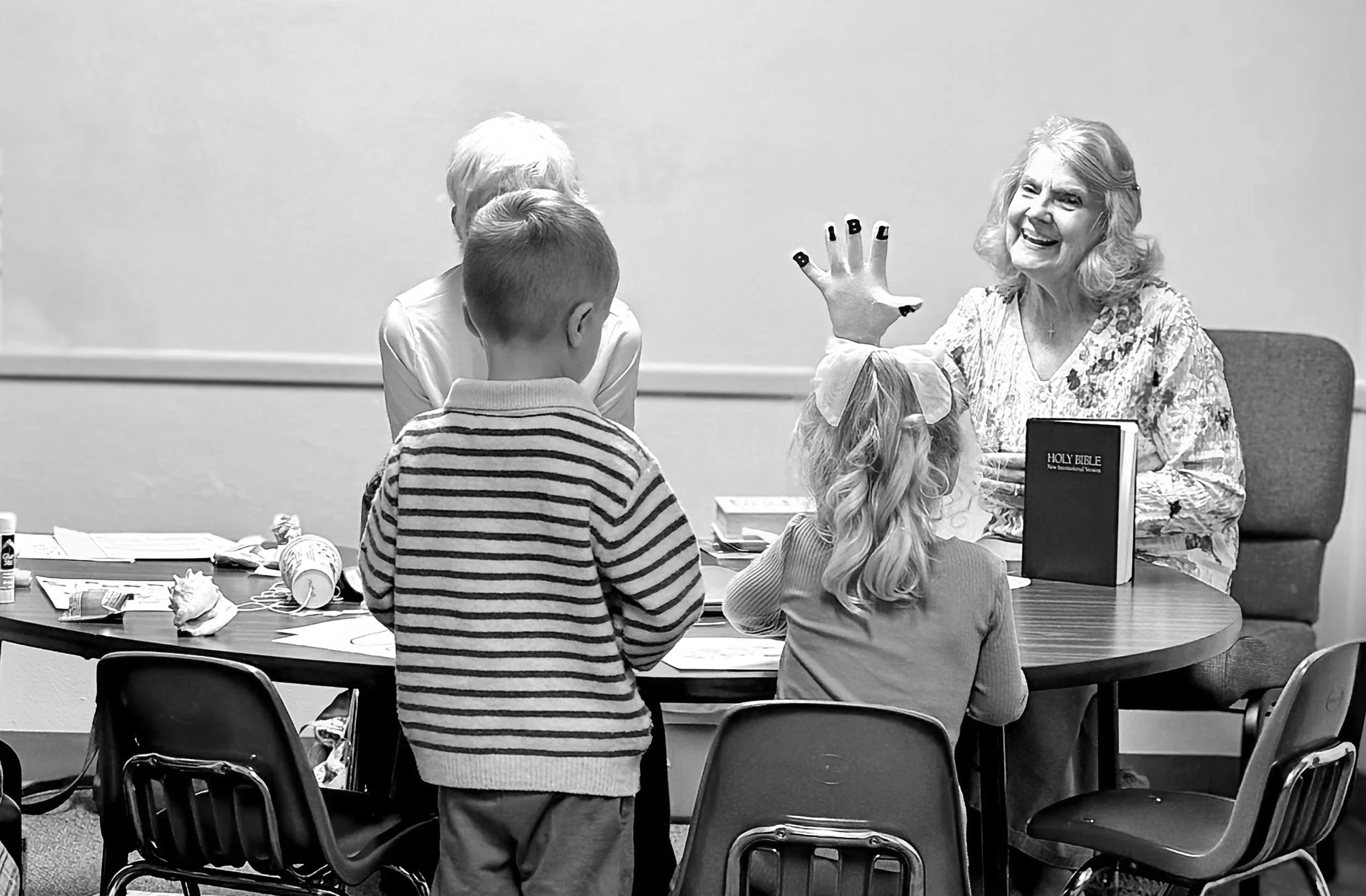 An elderly woman with gray hair, sitting at a table with children, holding her hand up with fingers labeled B I B L E, smiling as she teaches a Bible lesson to children, including a girl with a bow in her hair and a boy in a striped sweater.