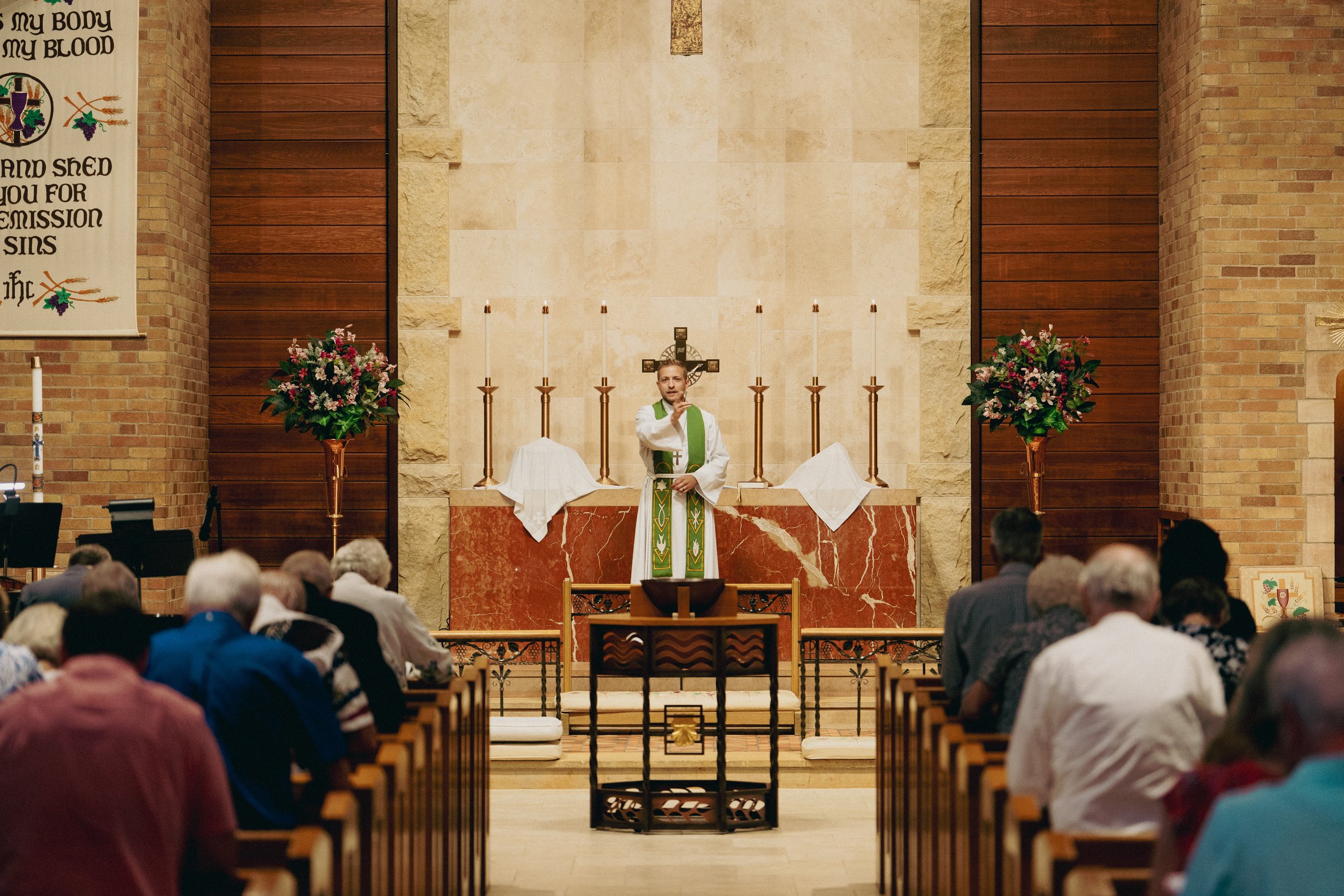 A pastor standing at the altar of a church. The altar has candles and floral arrangements. The church interior includes brick and wood walls, and there is a large crucifix behind the pastor.