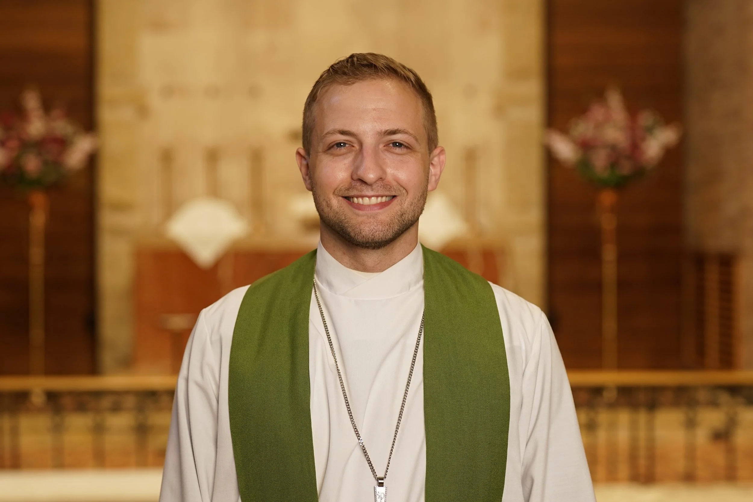 A smiling young man in a white religious robe and green stole standing inside a church with ornate decorations and flowers in the background.
